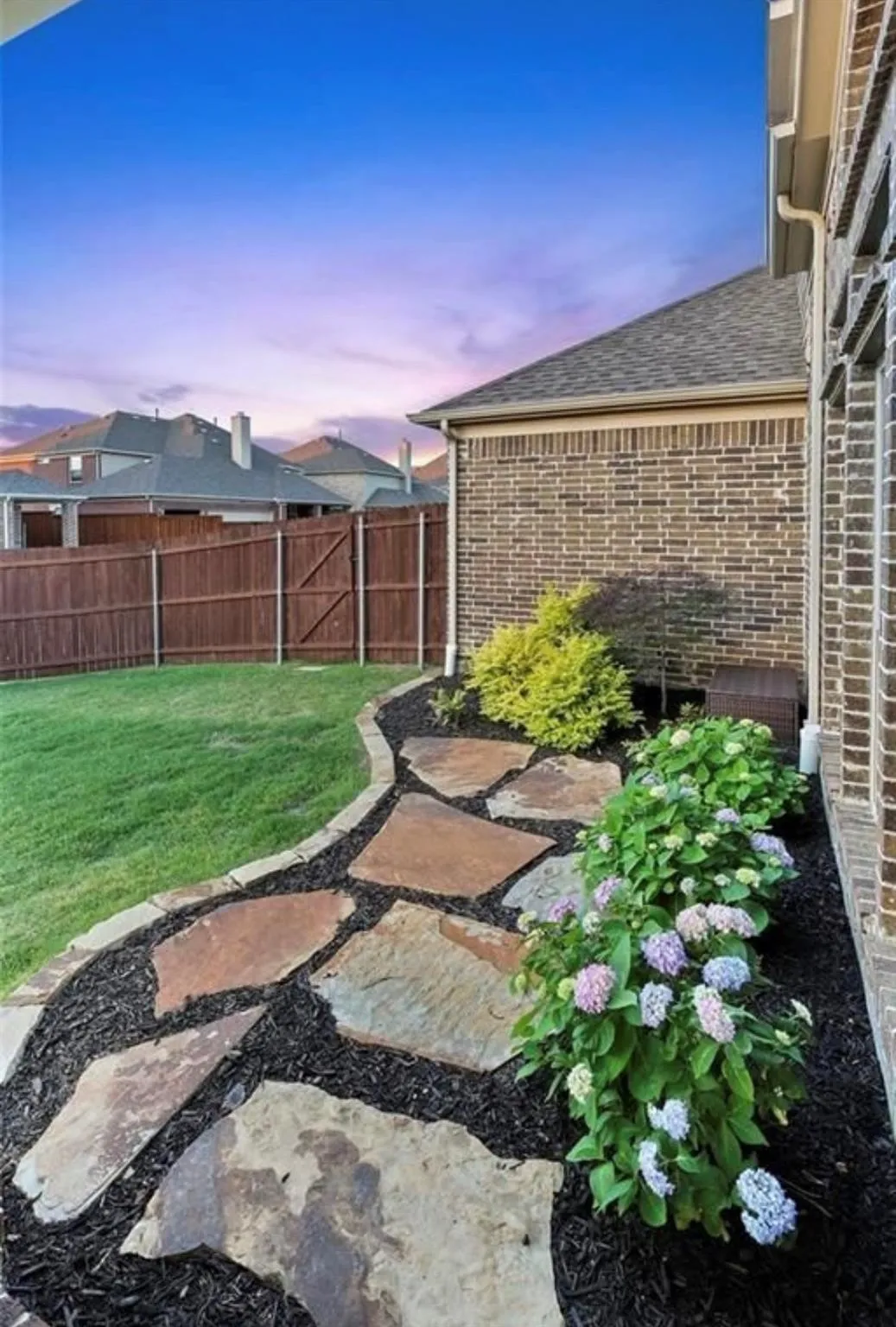 Side yard with flagstone pathway, mulched garden beds featuring established plantings, and a wooden privacy fence