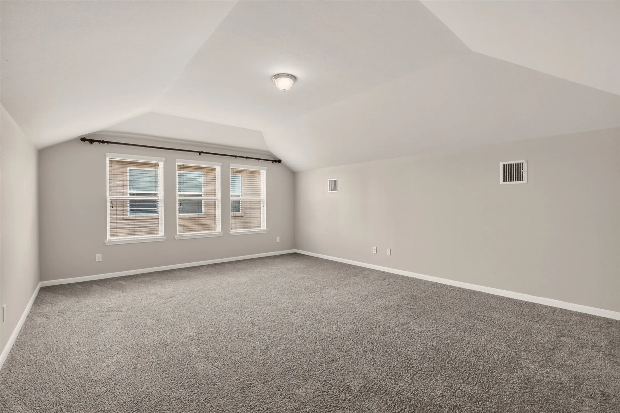 Spacious room featuring vaulted ceilings, three large windows with blinds, neutral carpeting, and light-toned wall paint