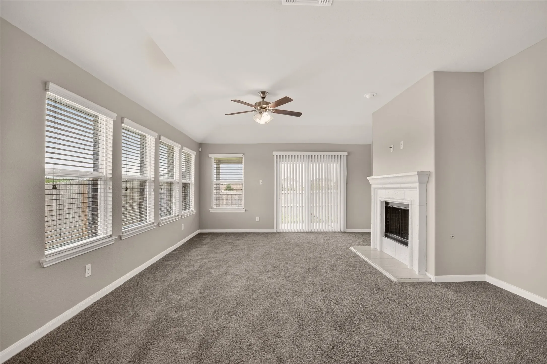 Spacious room featuring gray carpeting, a white mantel fireplace with a dark insert, a ceiling fan with integrated lighting, multiple windows with blinds, and a sliding glass door with vertical blinds