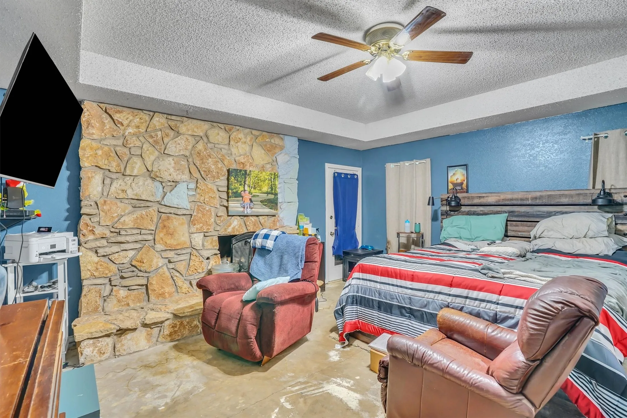 Spacious room featuring a stone-clad fireplace and a tray ceiling with a ceiling fan