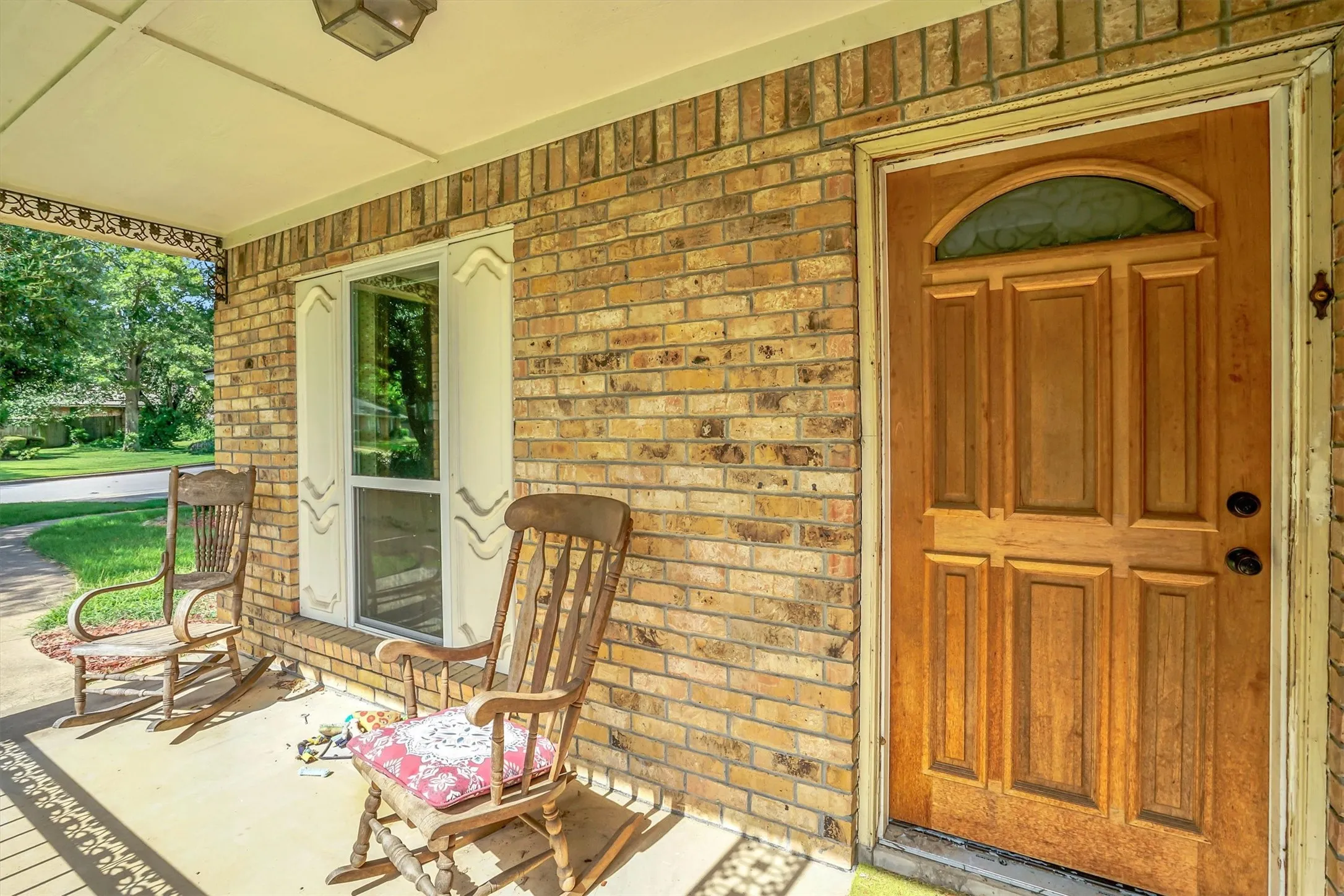 Brick exterior featuring a wood panel door with an arched glass insert, a covered porch, a single window with decorative shutters, and an outdoor ceiling light fixture