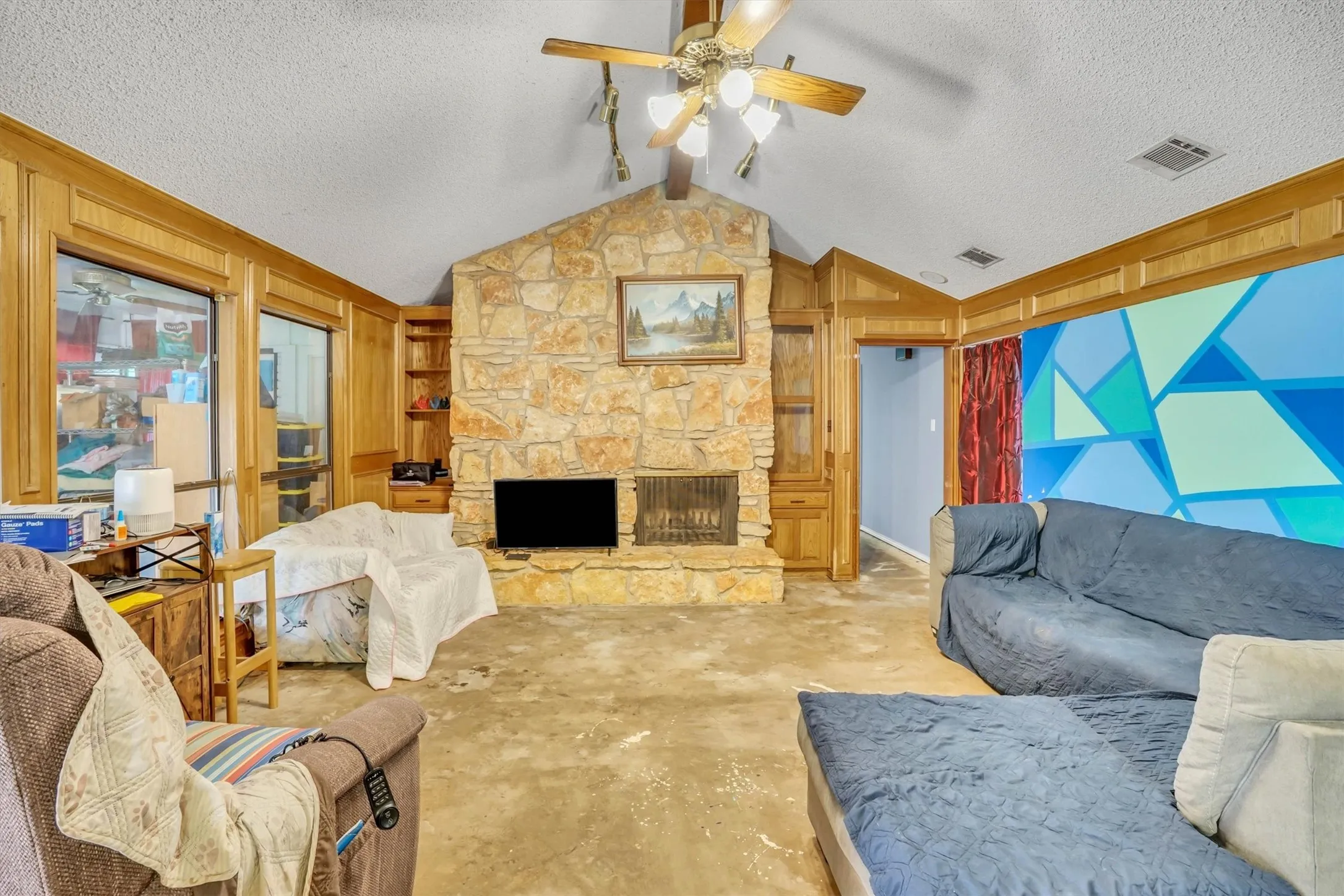 Vaulted ceiling living area featuring a prominent stone fireplace, built-in shelving, wood-paneled walls, and large windows