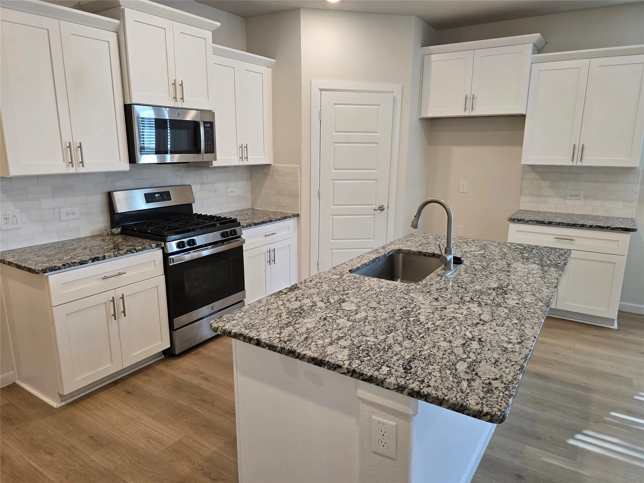 Kitchen featuring an island with an integrated sink, granite countertops, white shaker cabinetry with brushed nickel hardware, stainless steel appliances, and wood-finish flooring
