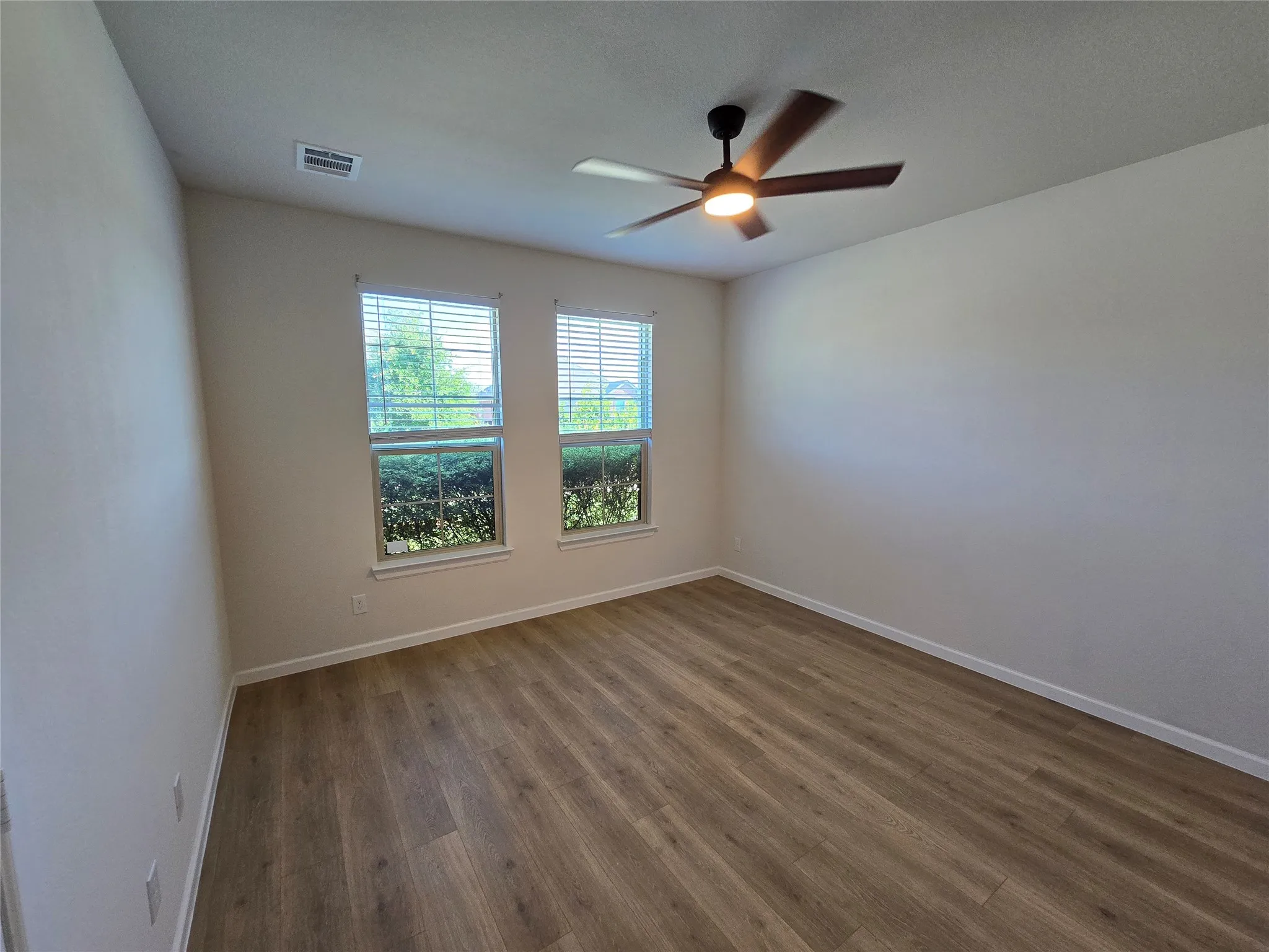 Spacious room with wood-finish flooring, two windows with blinds, a modern ceiling fan with integrated lighting, and neutral wall tones