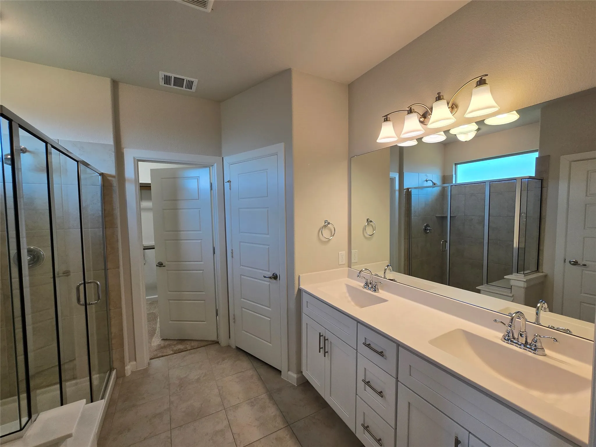 Spacious bathroom featuring a dual vanity with white cabinetry, a large mirror, a glass-enclosed shower, and tile flooring