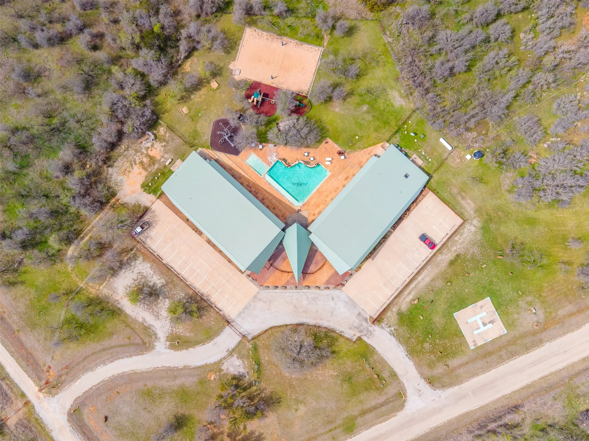 Aerial view showcasing a sprawling property featuring a central building with a light green roof, a swimming pool, extensive paved patio areas, and surrounding natural landscaping