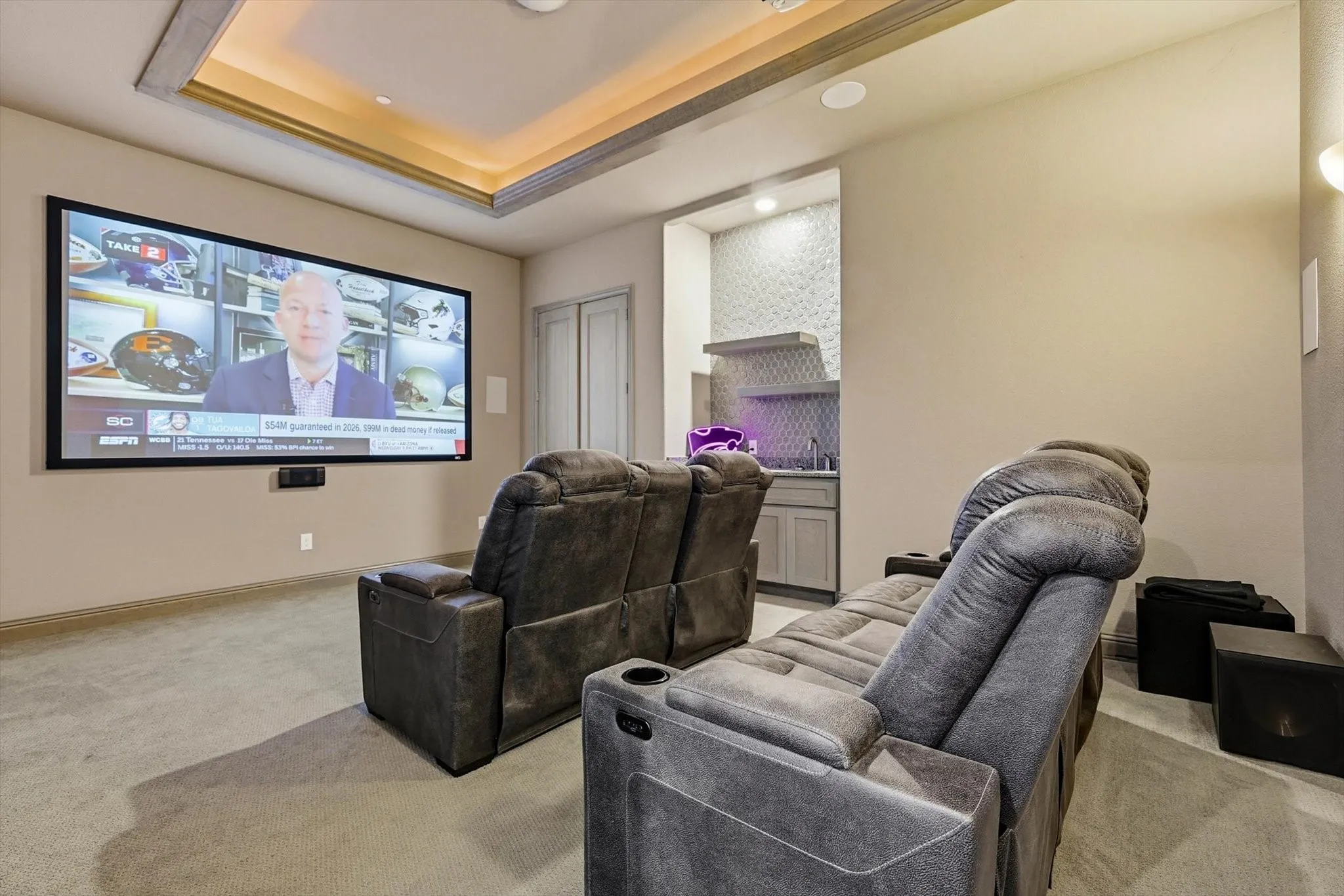 Dedicated media room featuring a large projection screen, recessed ceiling lighting, tray ceiling detail, built-in wet bar with textured tile backsplash, and neutral carpeting