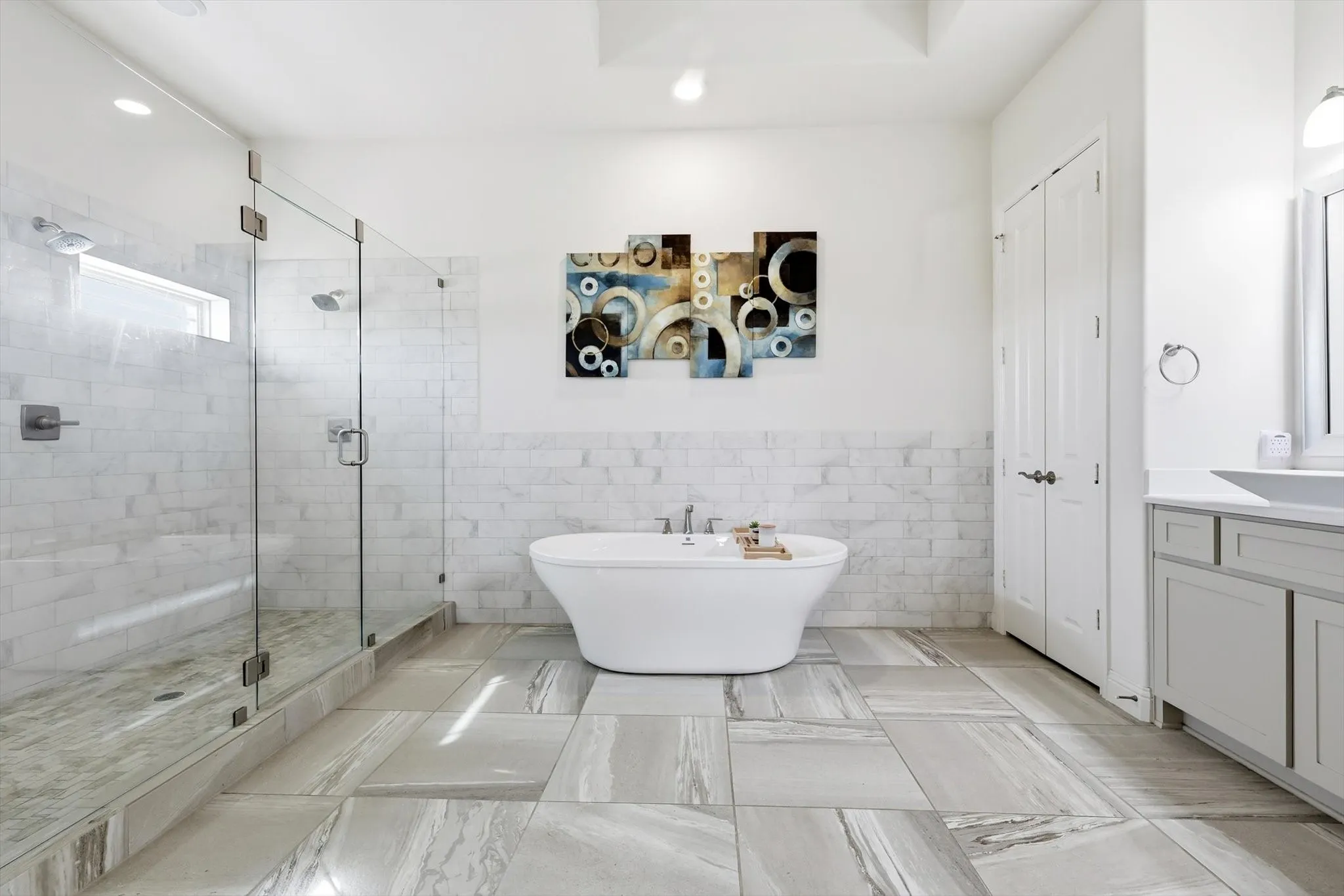 Modern bathroom featuring a freestanding soaking tub, a frameless glass shower enclosure with dual showerheads, light gray tile flooring, a single vanity with an undermount sink, and white shaker-style cabinetry