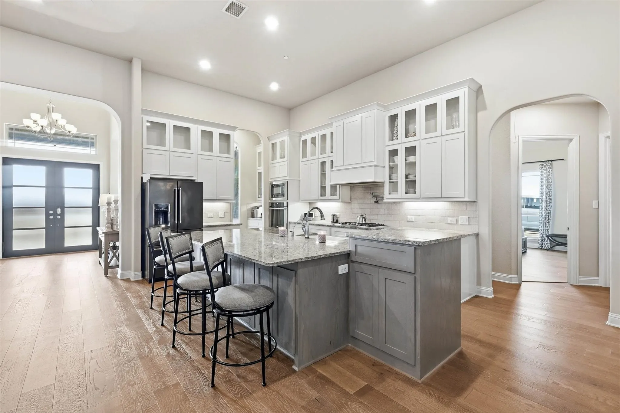 Open-concept kitchen featuring an oversized island with seating, light wood-finish flooring, white upper cabinetry with glass fronts, and a subway tile backsplash