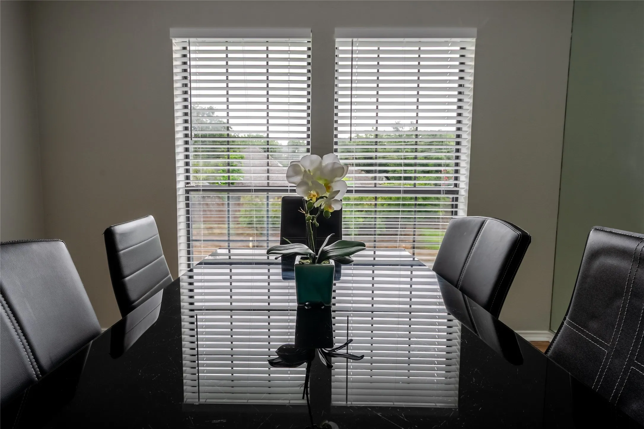 Dining area featuring a dark, reflective surface table and two large windows with horizontal blinds