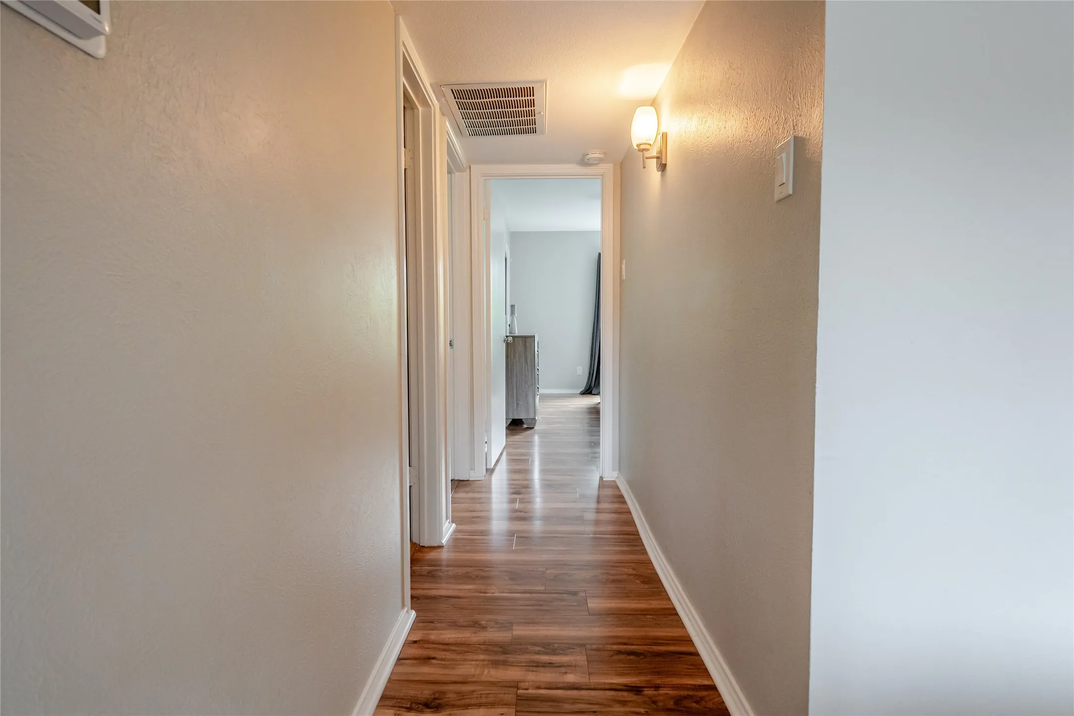 Hallway featuring wood-finish flooring, light grey walls, white baseboards, and a wall-mounted sconce