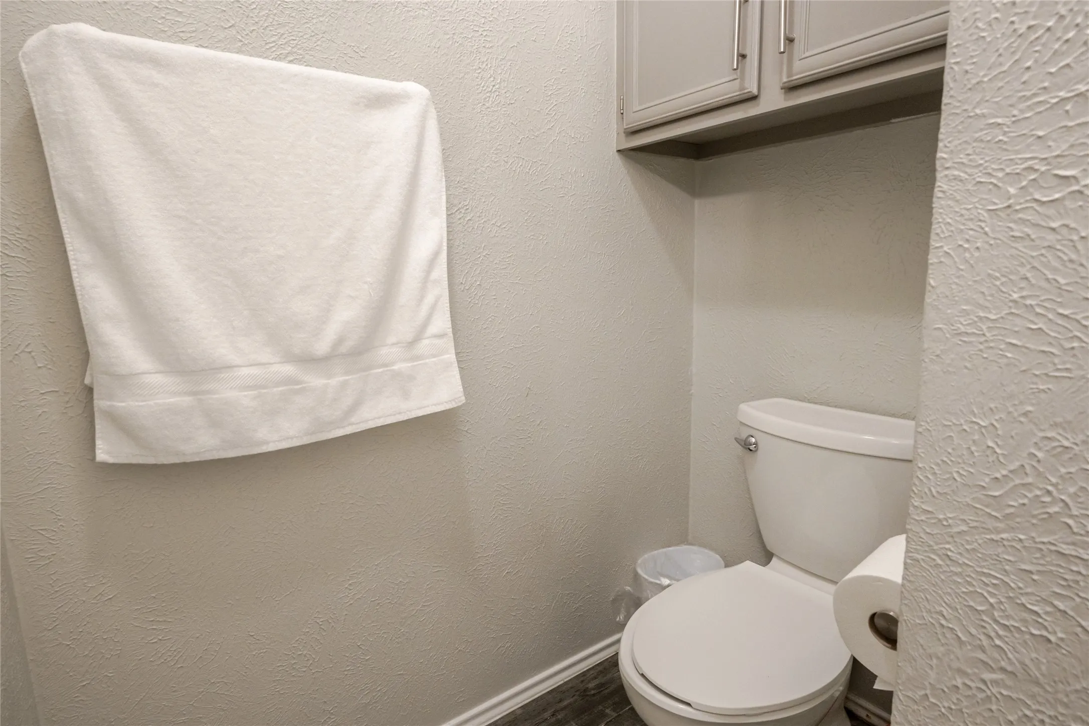 Compact water closet featuring a white porcelain toilet, overhead cabinetry, and wood-finish flooring