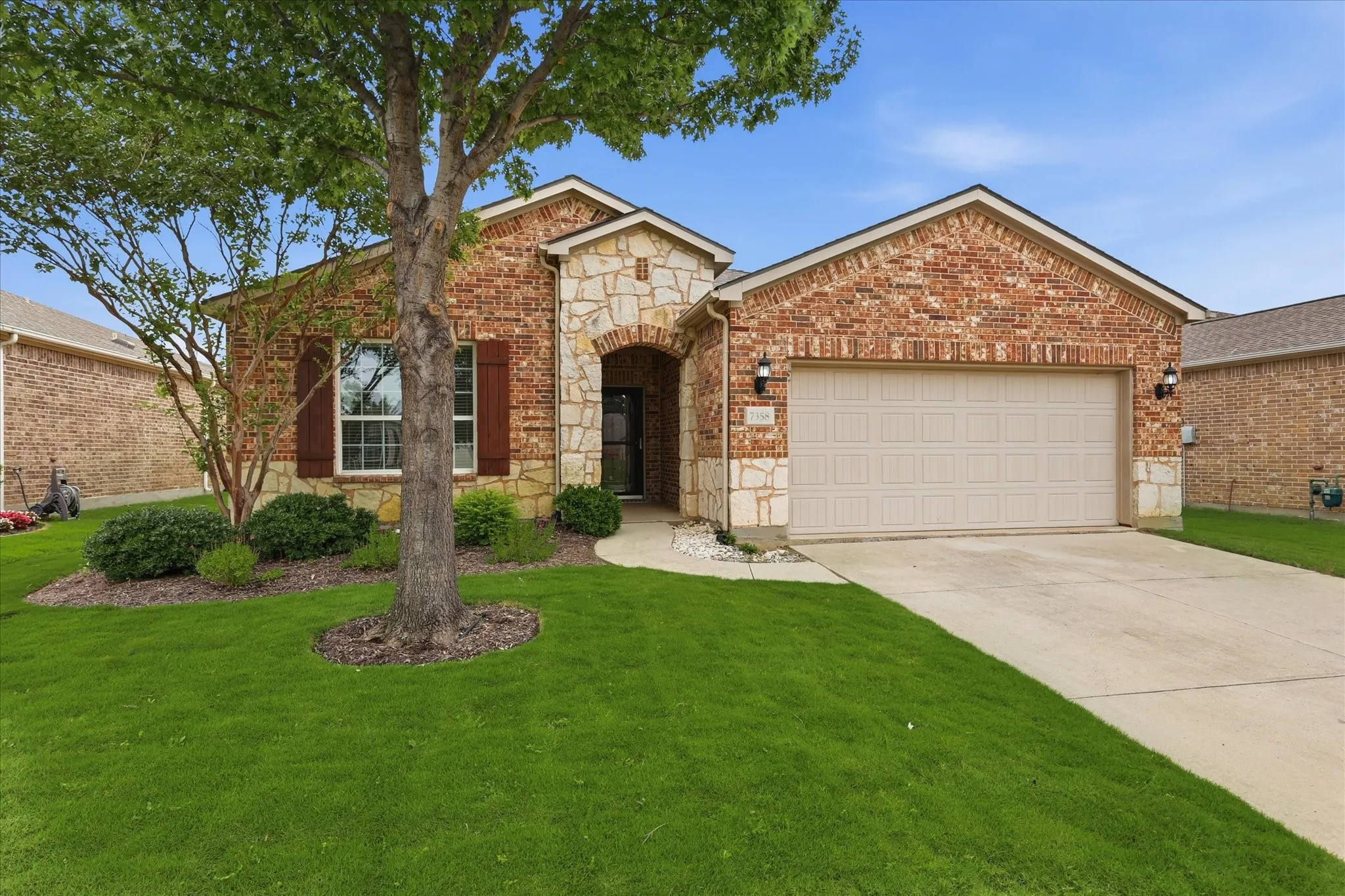 Brick and stone facade residence featuring a two-car garage, mature trees, manicured lawn, and a covered entry