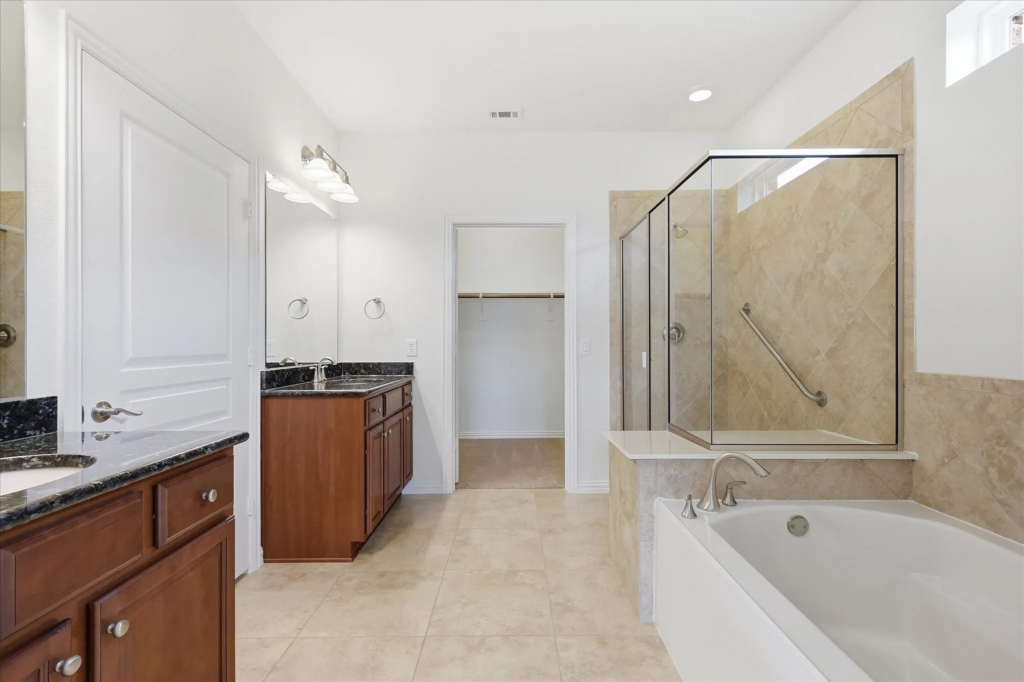 Spacious bathroom featuring dual wood-finish vanities with dark stone countertops, a large soaking tub with deck-mounted fixtures, and a clear glass shower enclosure with tile surround