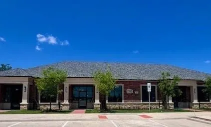 Brick and stone facade building with large window displays, columned entryways, a gray shingle roof, and ample front-facing parking