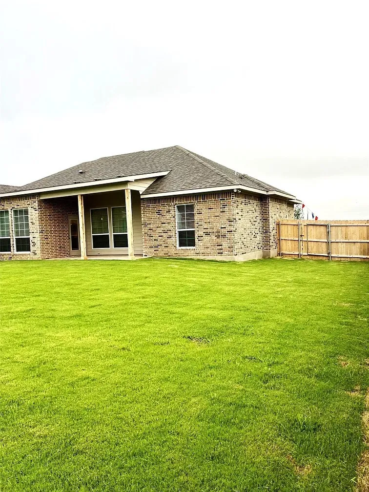 Expansive green lawn with a covered patio, brick exterior, multiple windows, and a wood privacy fence