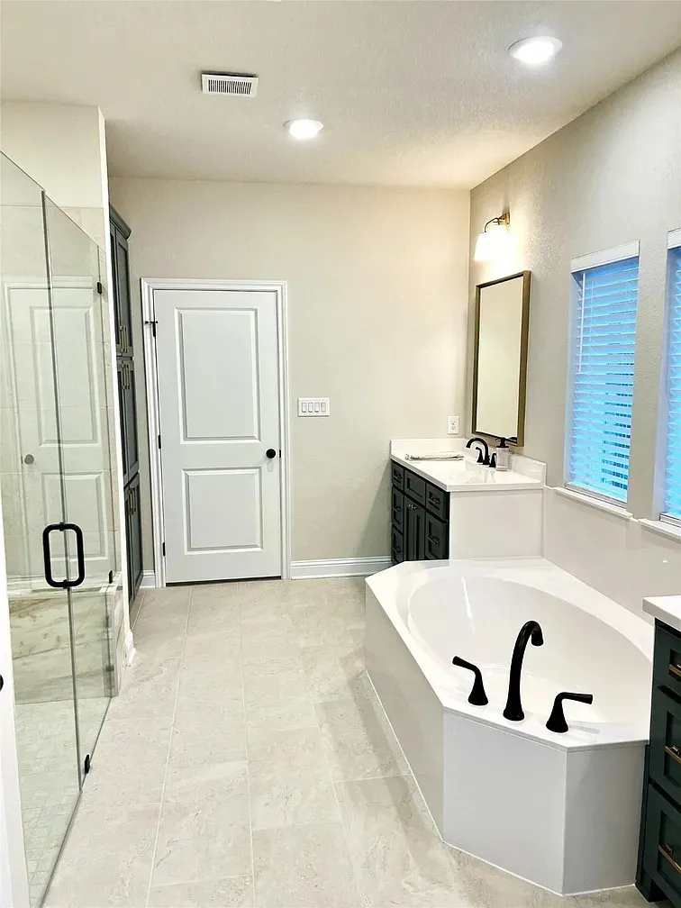 Spacious bathroom featuring a white soaking tub with black fixtures, a walk-in shower with glass enclosure, dual dark cabinetry vanities with white countertops, and light tile flooring throughout