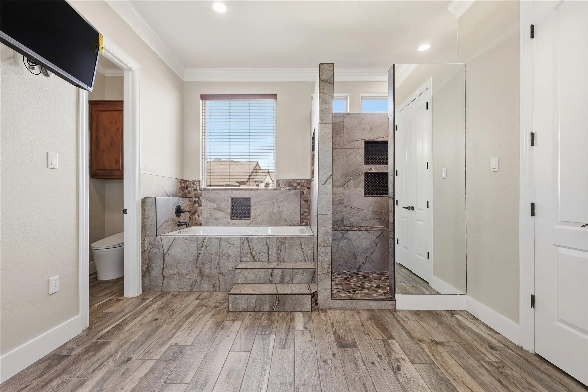 Spacious bathroom featuring wood-finish flooring, a tiled soaking tub with steps, a walk-in shower with stone pebble flooring and built-in shelving, and a separate water closet