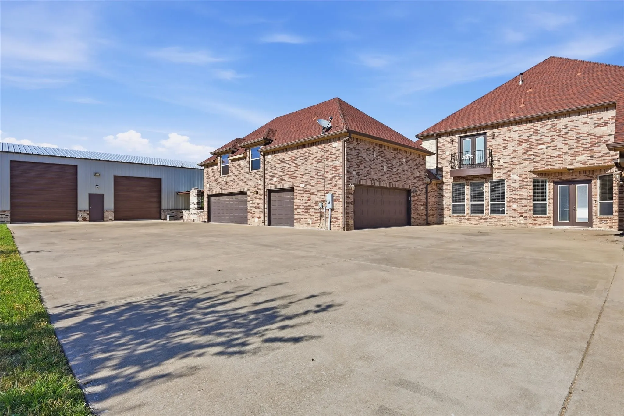 Expansive concrete driveway with a large brick structure featuring two garage doors and a balcony, alongside a metal-sided building with two additional garage doors and a pedestrian door