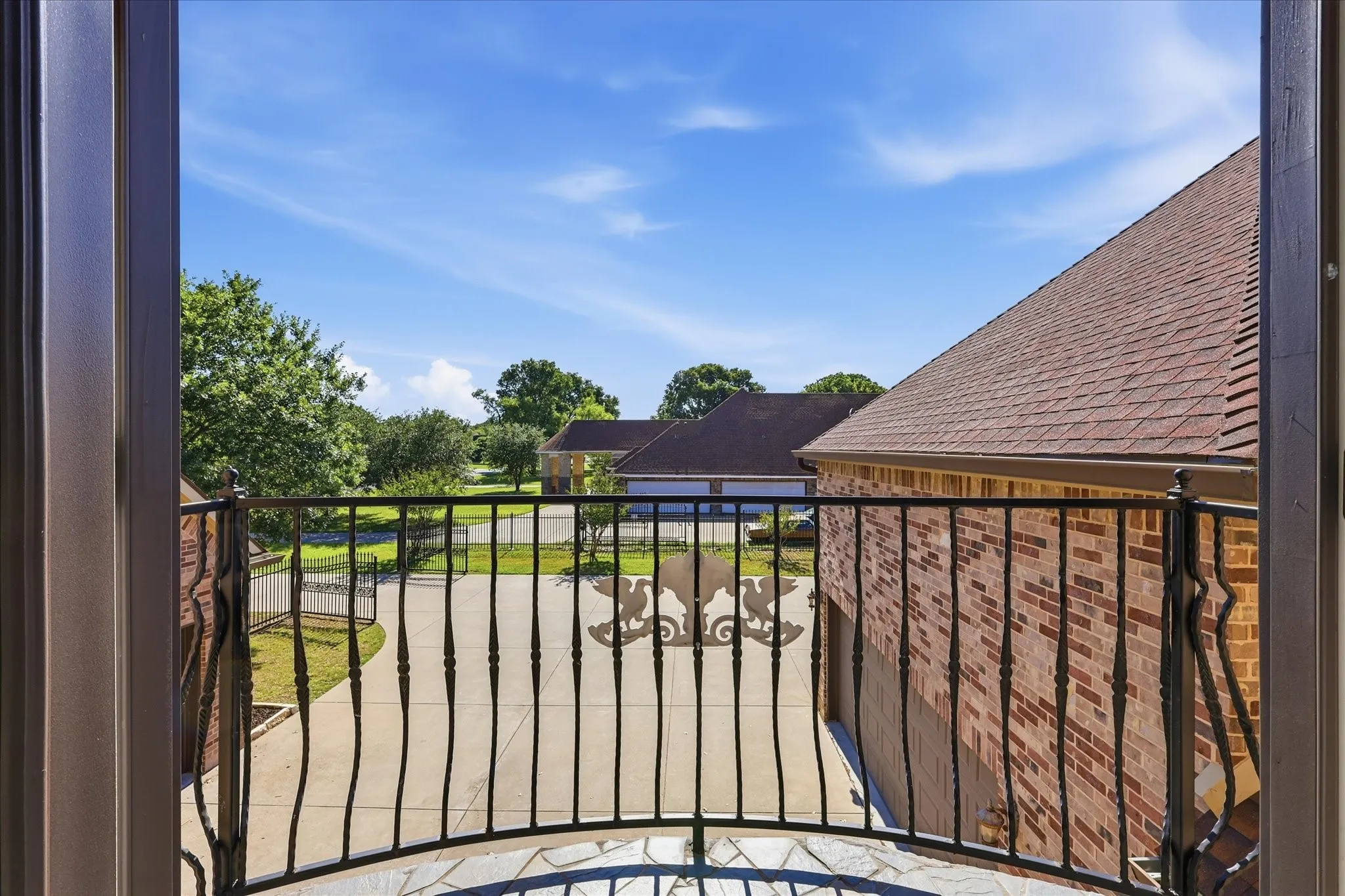 Balcony featuring an ornate wrought iron railing, stone tile flooring, and a view of the concrete driveway and brick exterior