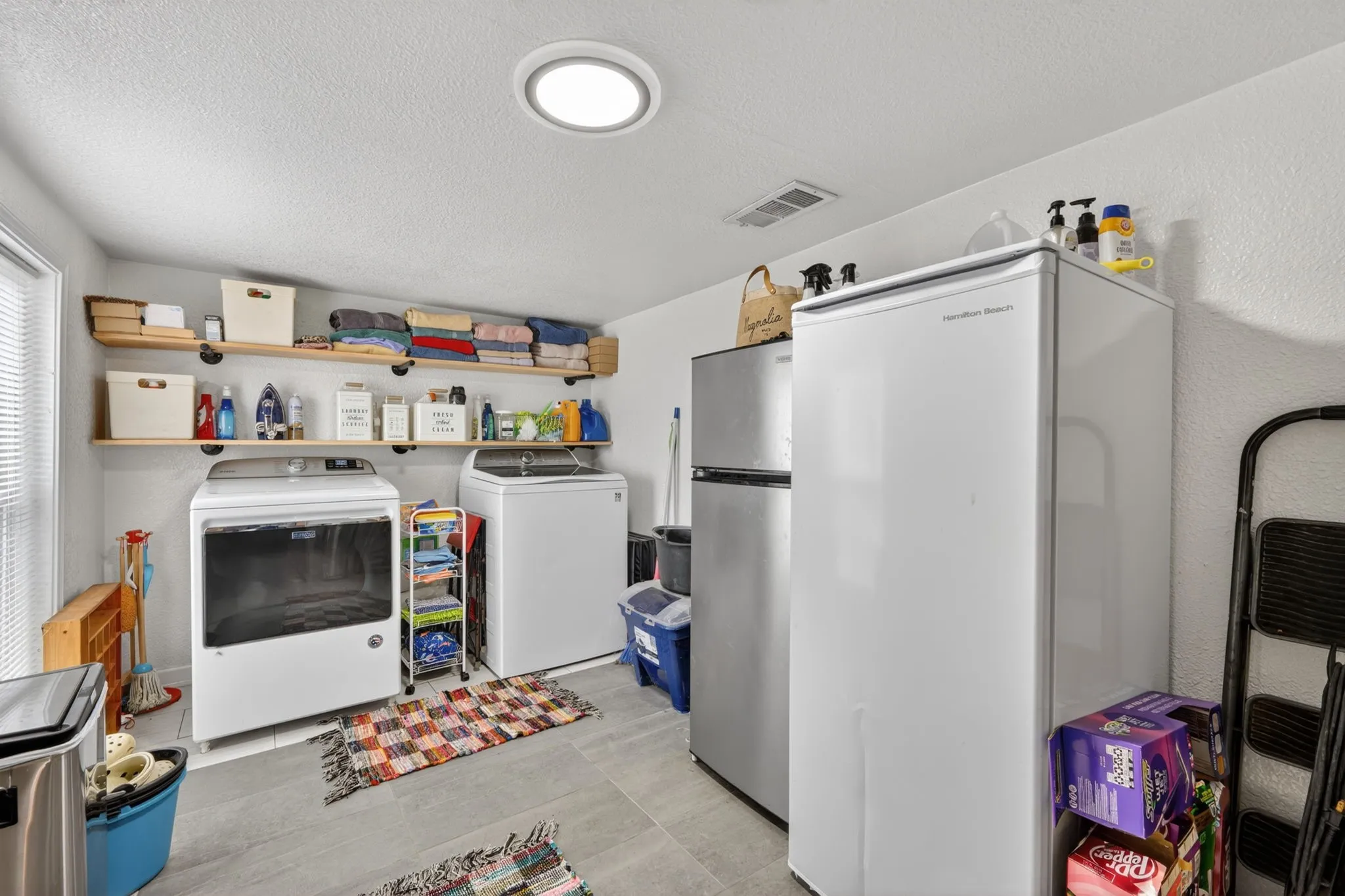 Dedicated utility space featuring light-toned flooring, overhead shelving, and a prominent window with horizontal blinds