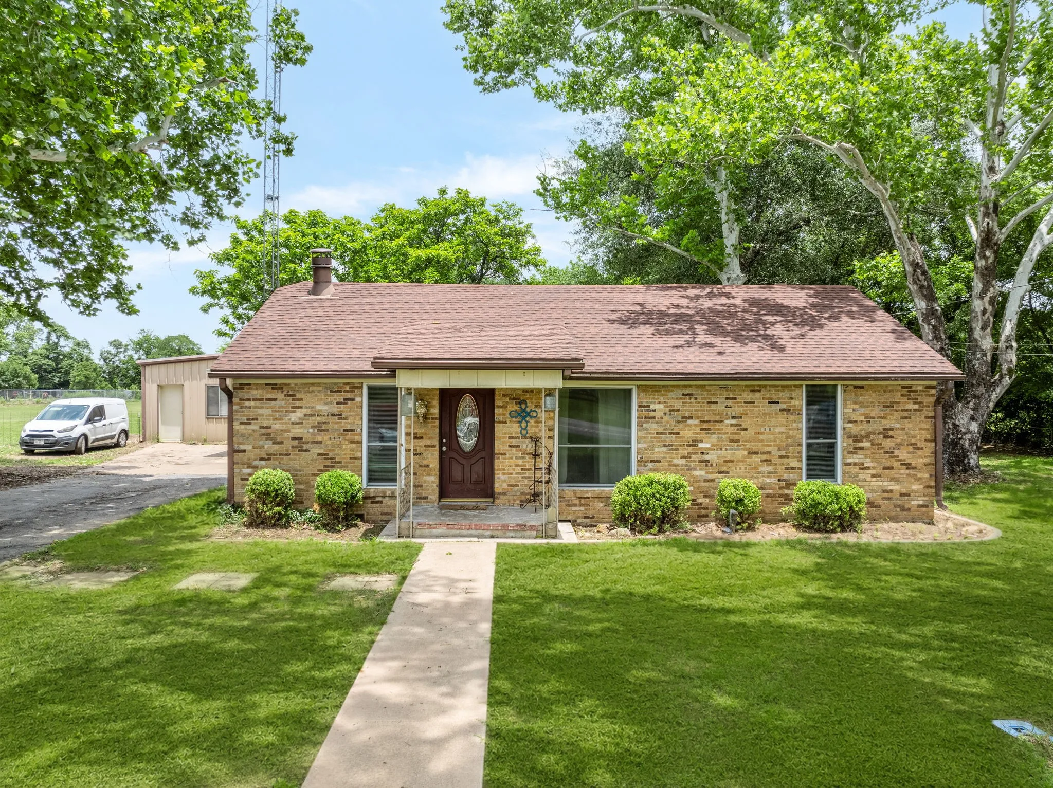 Brick exterior residence featuring a front porch entry, a concrete walkway, mature trees, and a detached metal building