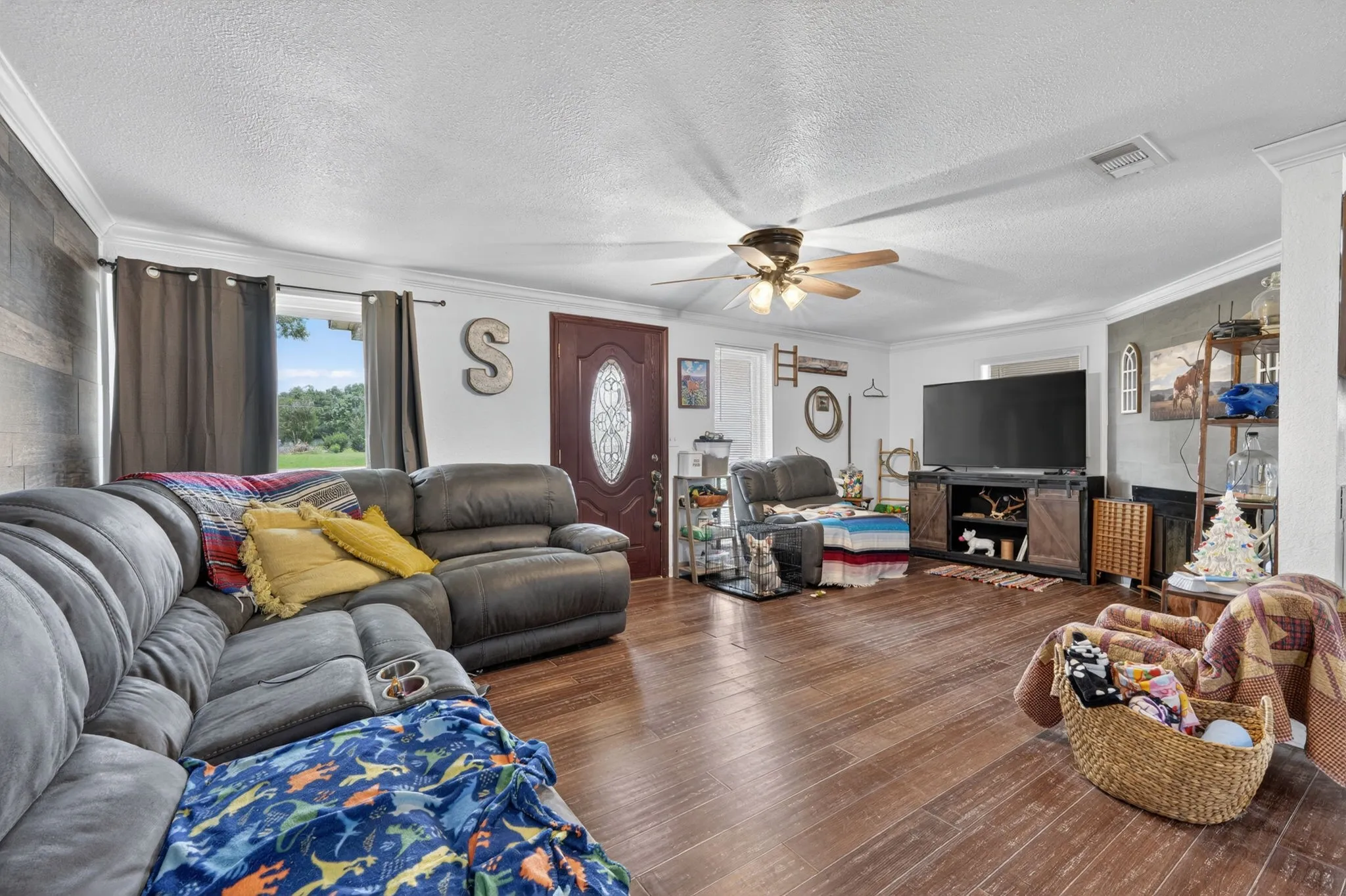Spacious living area featuring wood-finish flooring, a decorative accent wall, a ceiling fan with integrated lighting, a front entry door with an oval glass insert, and crown molding