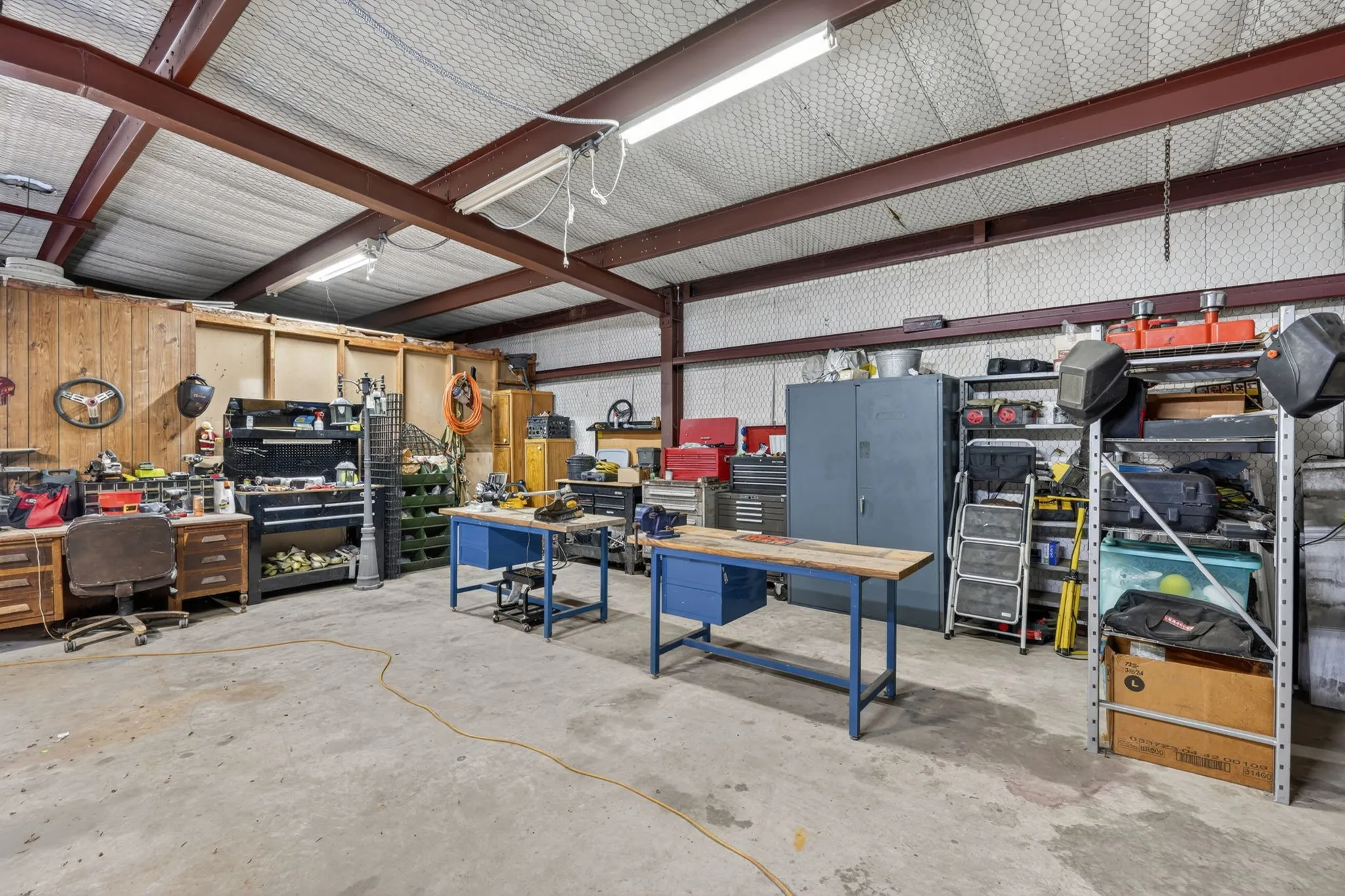 Spacious workshop featuring exposed structural beams, high ceilings, concrete flooring, and ample workspace with built-in wooden workbenches