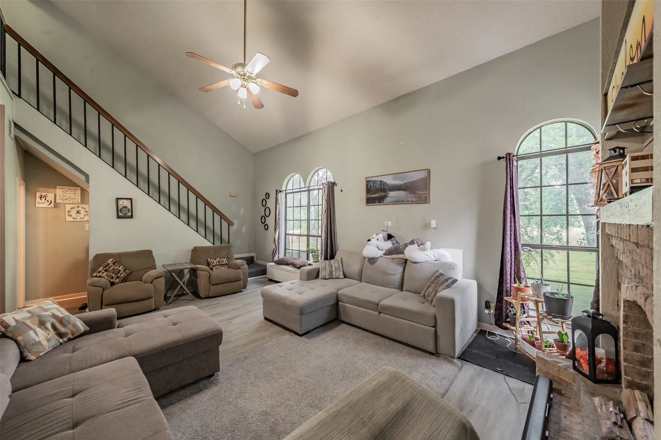 Spacious living area featuring a vaulted ceiling with a ceiling fan, a prominent staircase with black metal balusters and wood handrails, and a fireplace with a brick surround