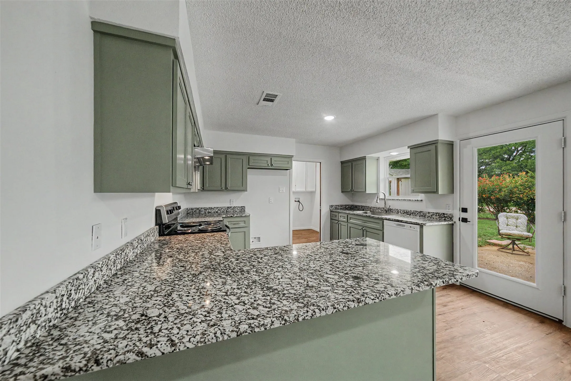 Kitchen featuring ample cabinetry in a muted green finish, speckled countertops, wood-finish flooring, recessed lighting, and a white paneled door providing outdoor access