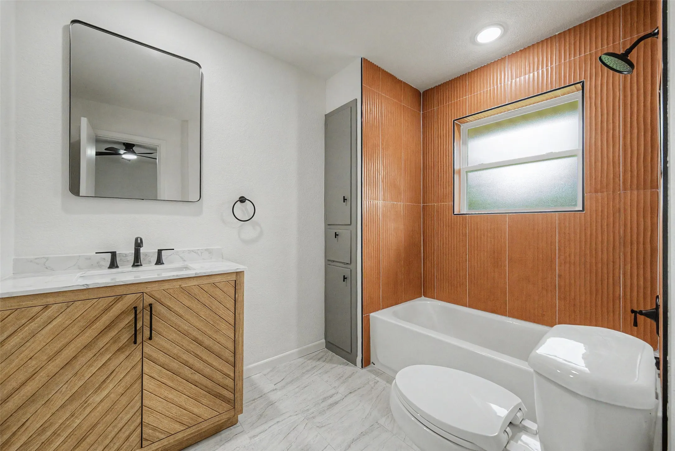 Bathroom featuring an orange-toned textured tile shower surround, a built-in bathtub, and a wood-finish vanity with a marble-patterned countertop