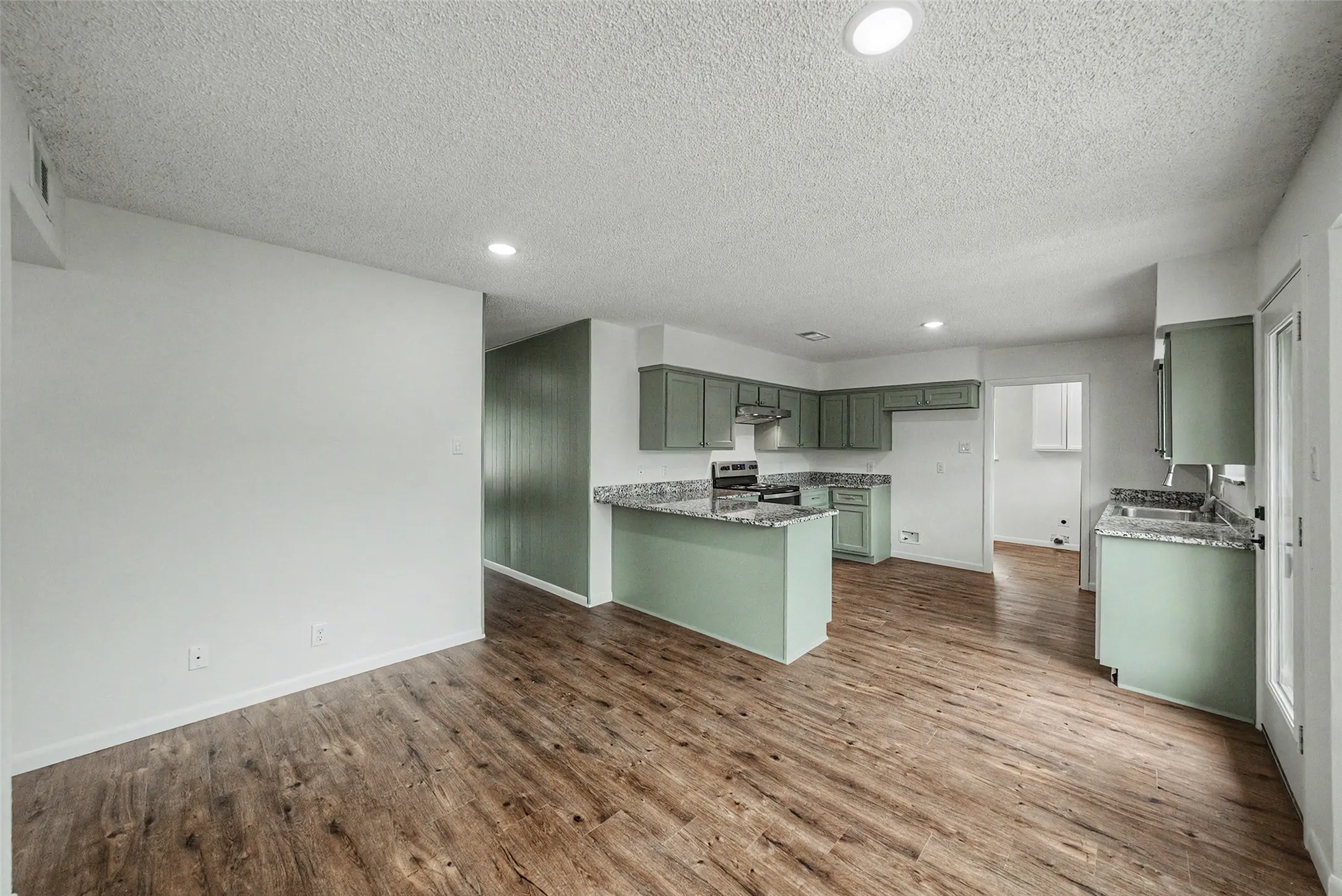 Open-concept kitchen and living area featuring wood-finish flooring, recessed lighting, sage green cabinetry, and granite-style countertops
