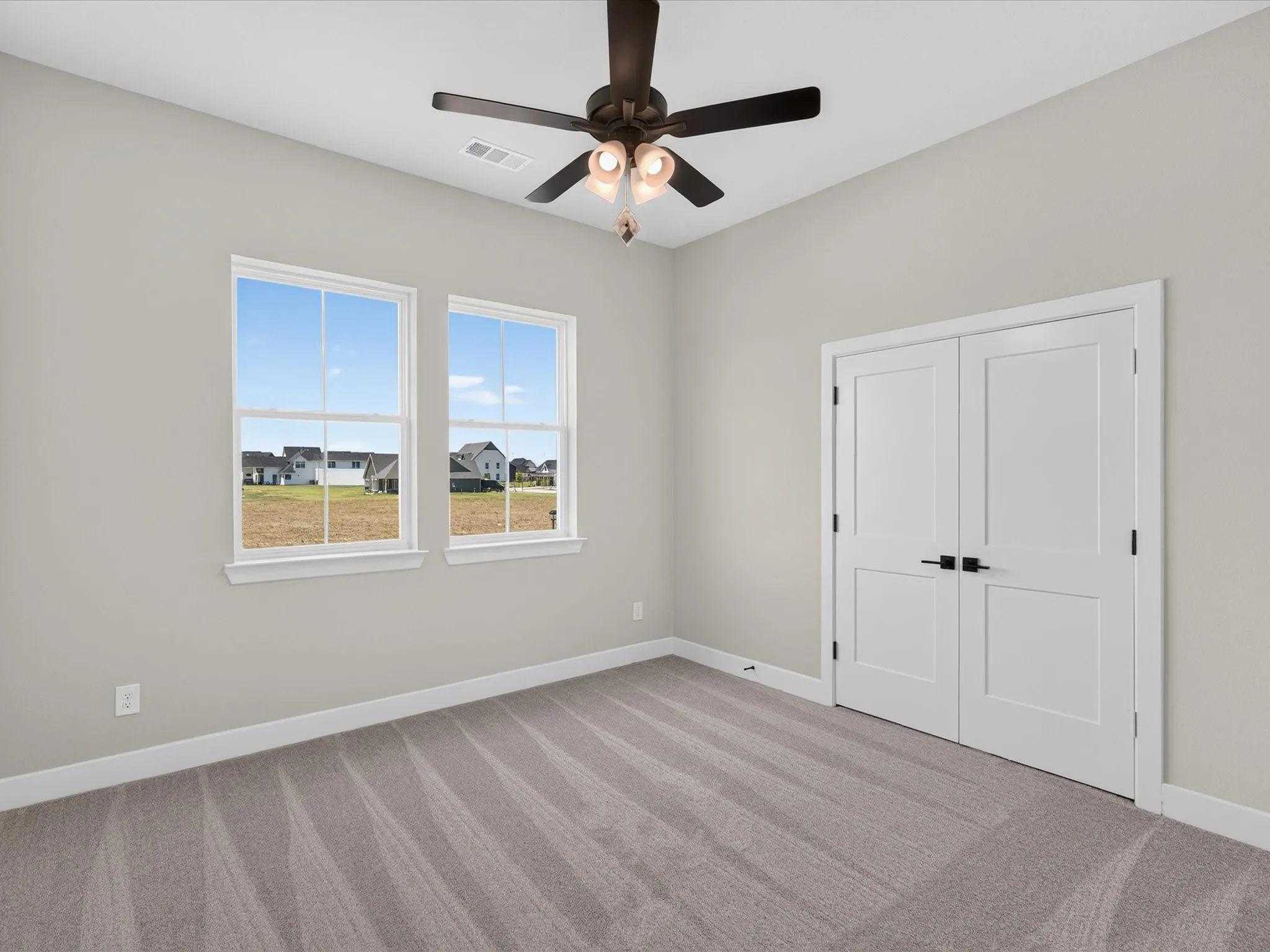 Neutral wall paint, two double-hung windows, carpeted flooring, white bi-fold doors with black hardware, and a ceiling fan with integrated lighting