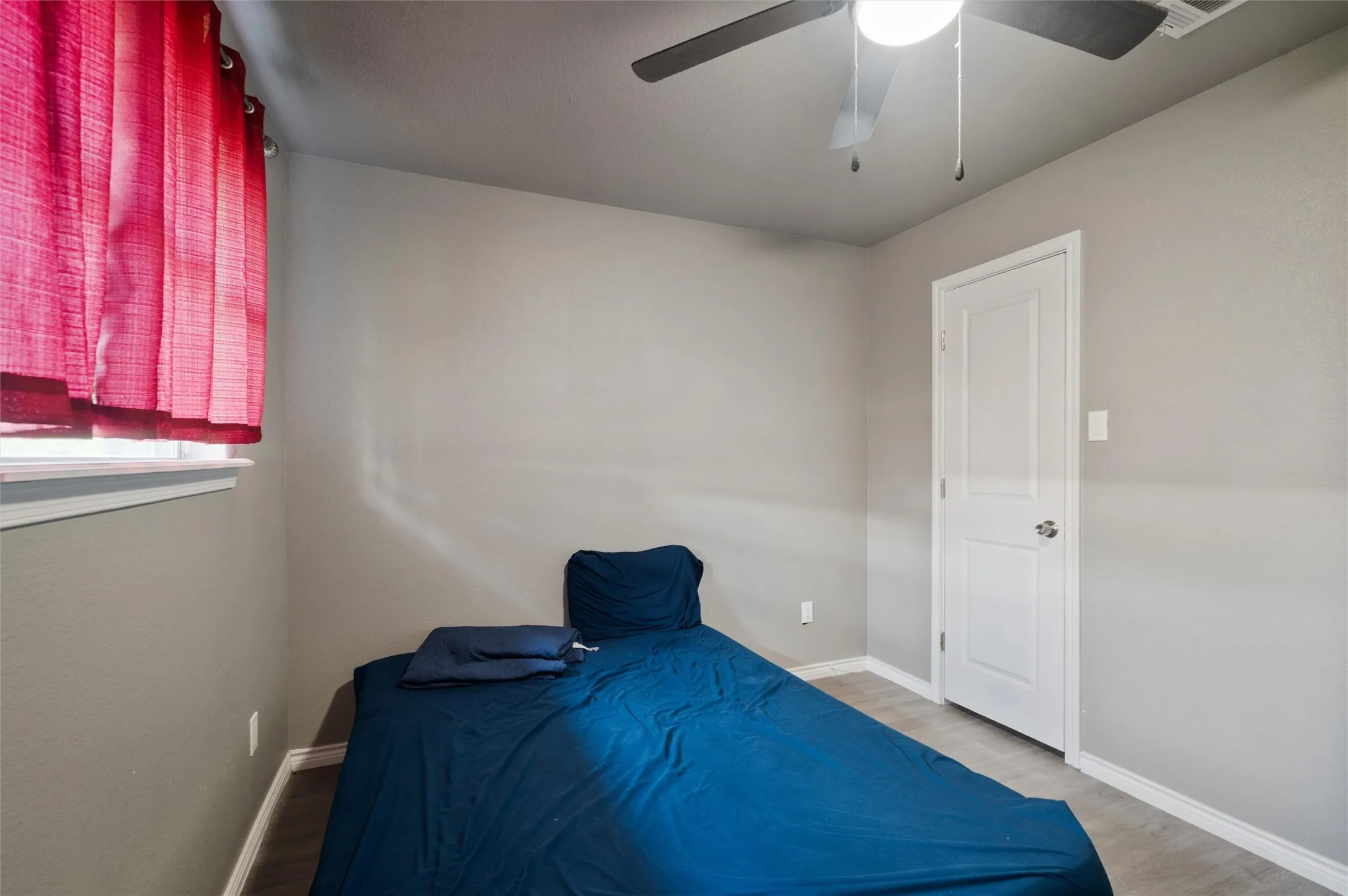 Light gray painted walls with white trim, wood-finish flooring, a white paneled door, a window with red blinds, and a ceiling fan with integrated lighting