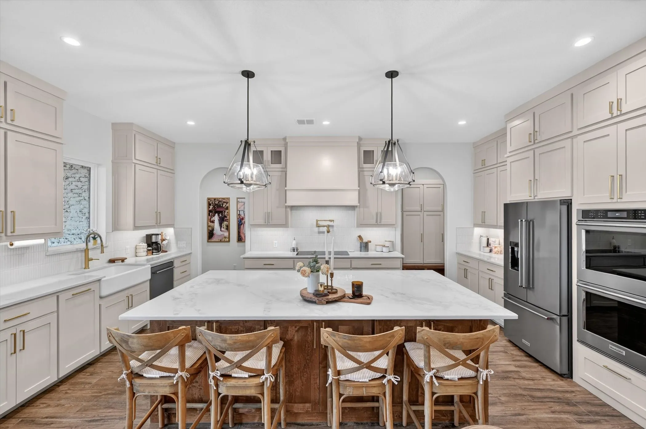 Expansive kitchen featuring a large central island with a light-toned countertop, wood-finish flooring, full-height cabinetry with gold-toned hardware, stainless steel appliances, and recessed lighting