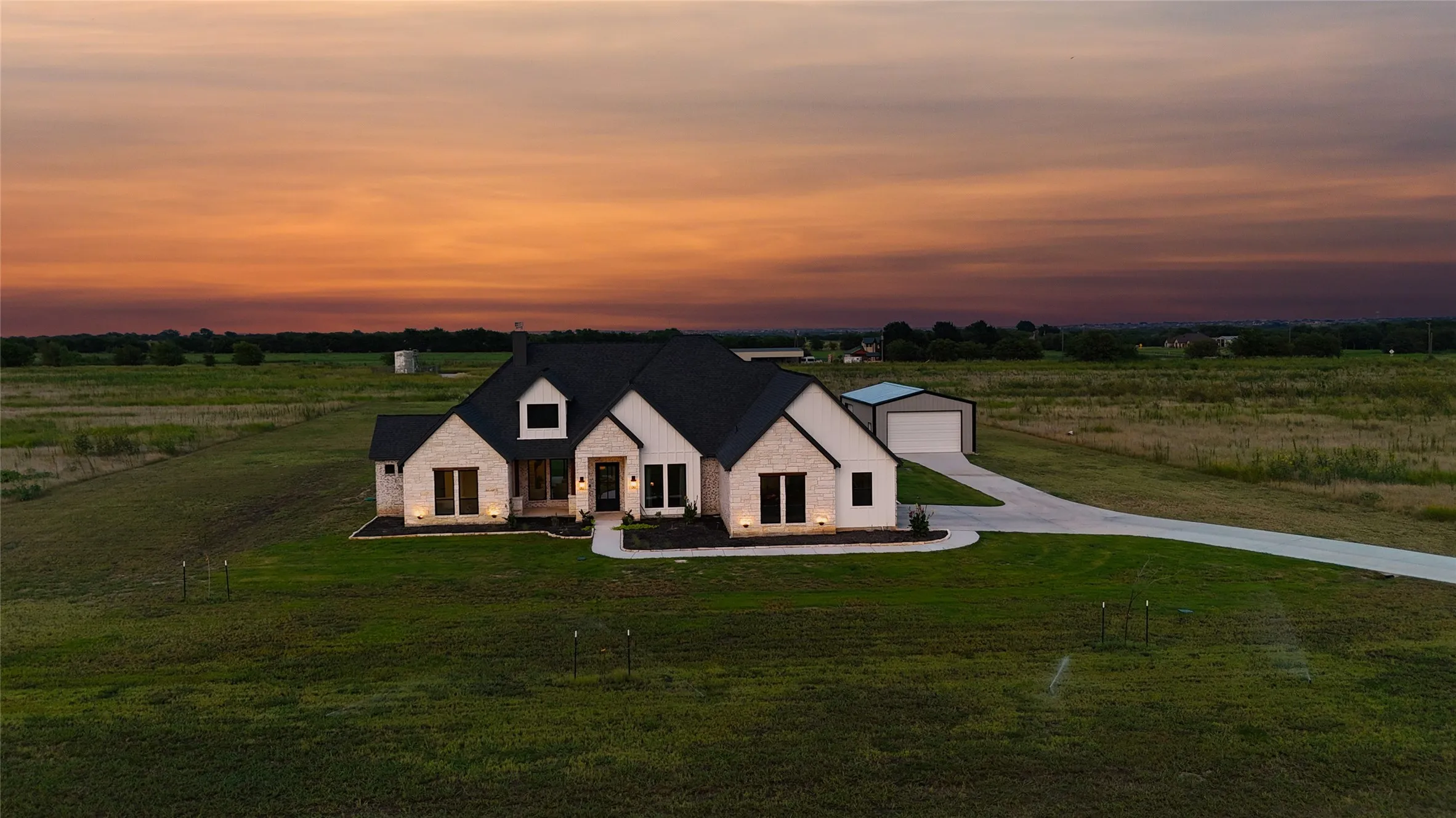 White stone exterior with dark trim and black roof, attached two-car garage, separate outbuilding with garage door, concrete driveway, and expansive green lawn