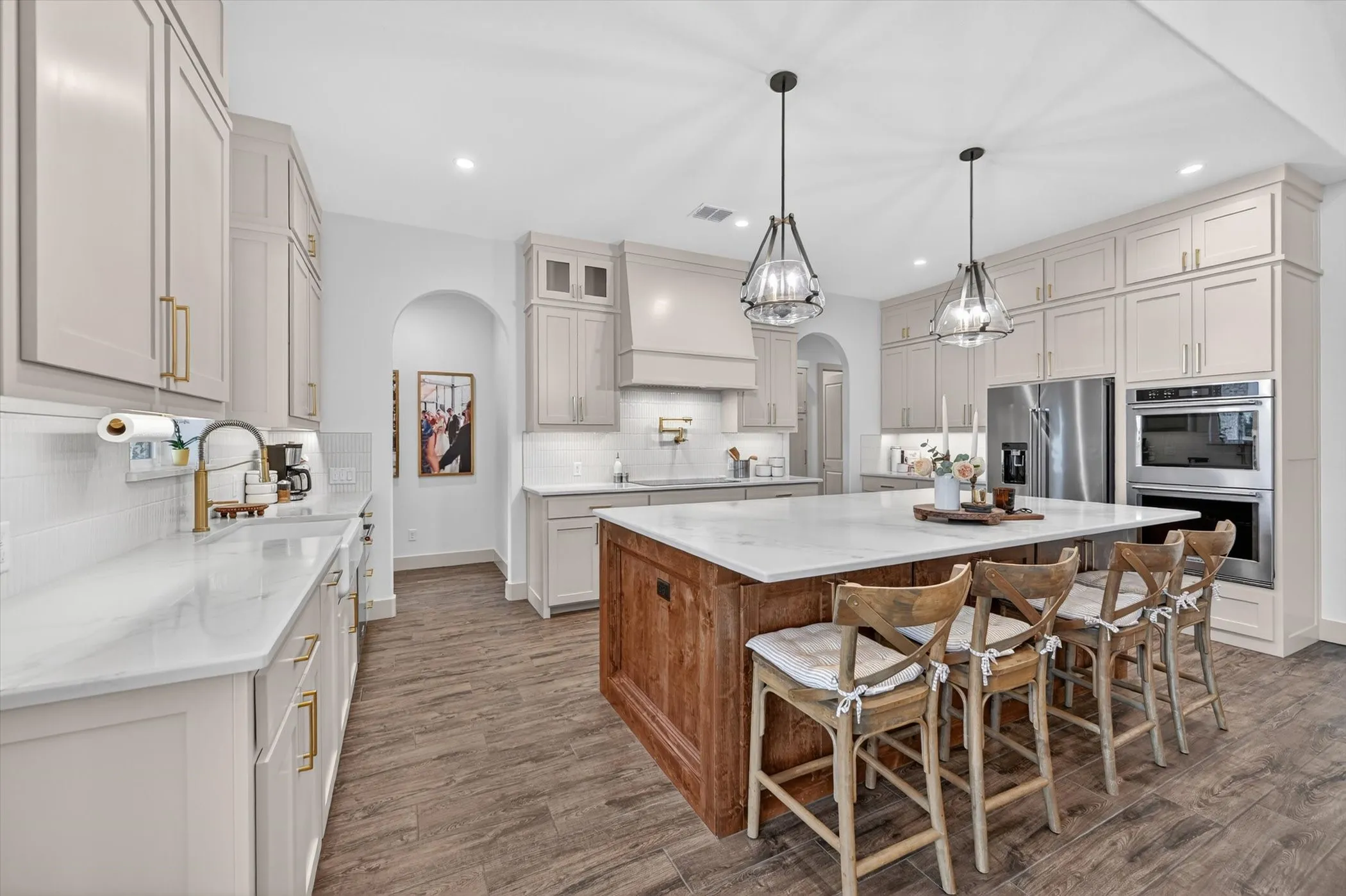 Expansive kitchen featuring a large central island with a stone countertop, extensive light-beige cabinetry, stainless steel appliances, wood-look tile flooring, and pendant lighting