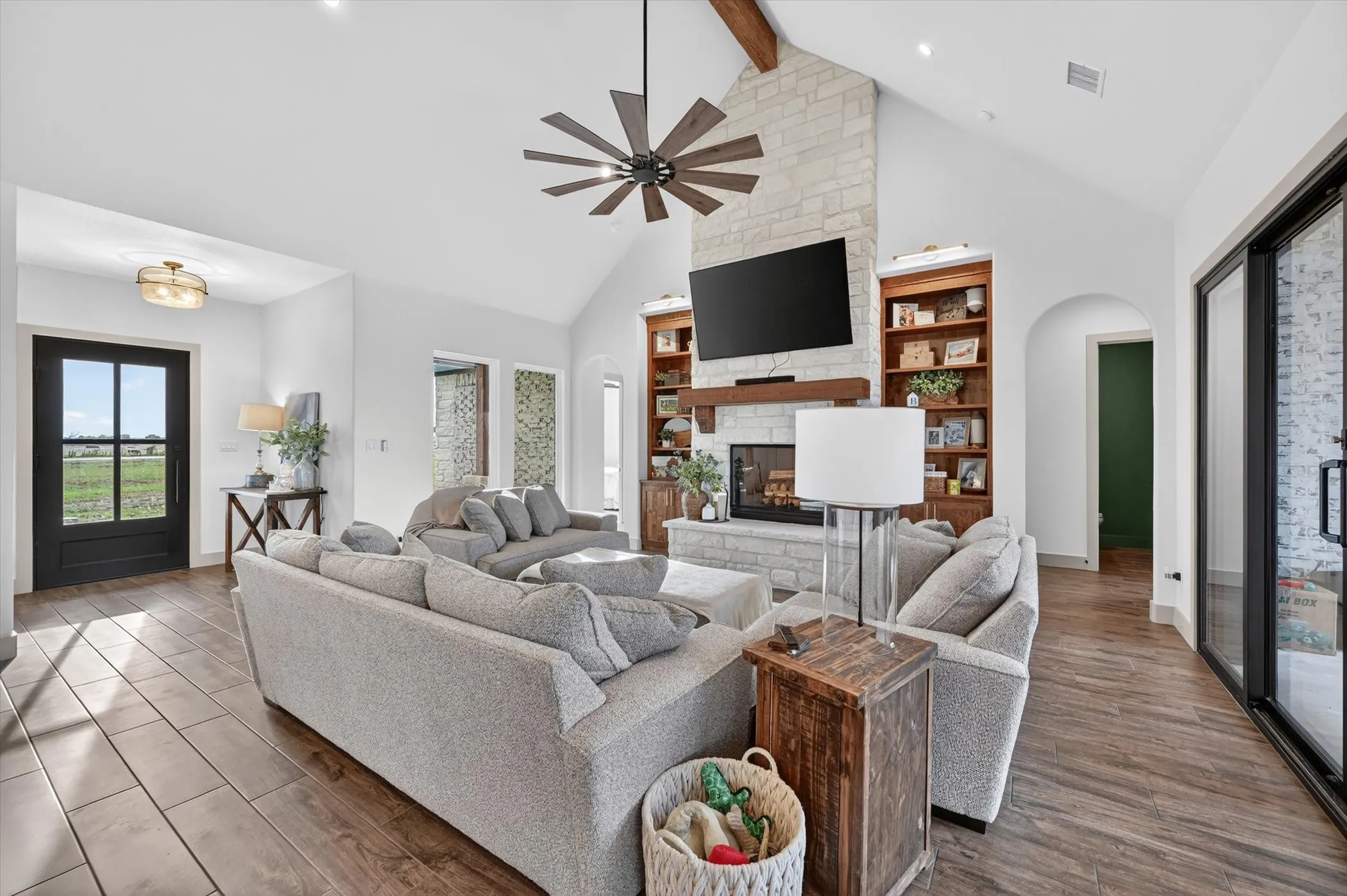 Vaulted living space featuring a stone-clad fireplace with built-in shelving, wood-look tile flooring, a contemporary ceiling fan, and abundant natural light
