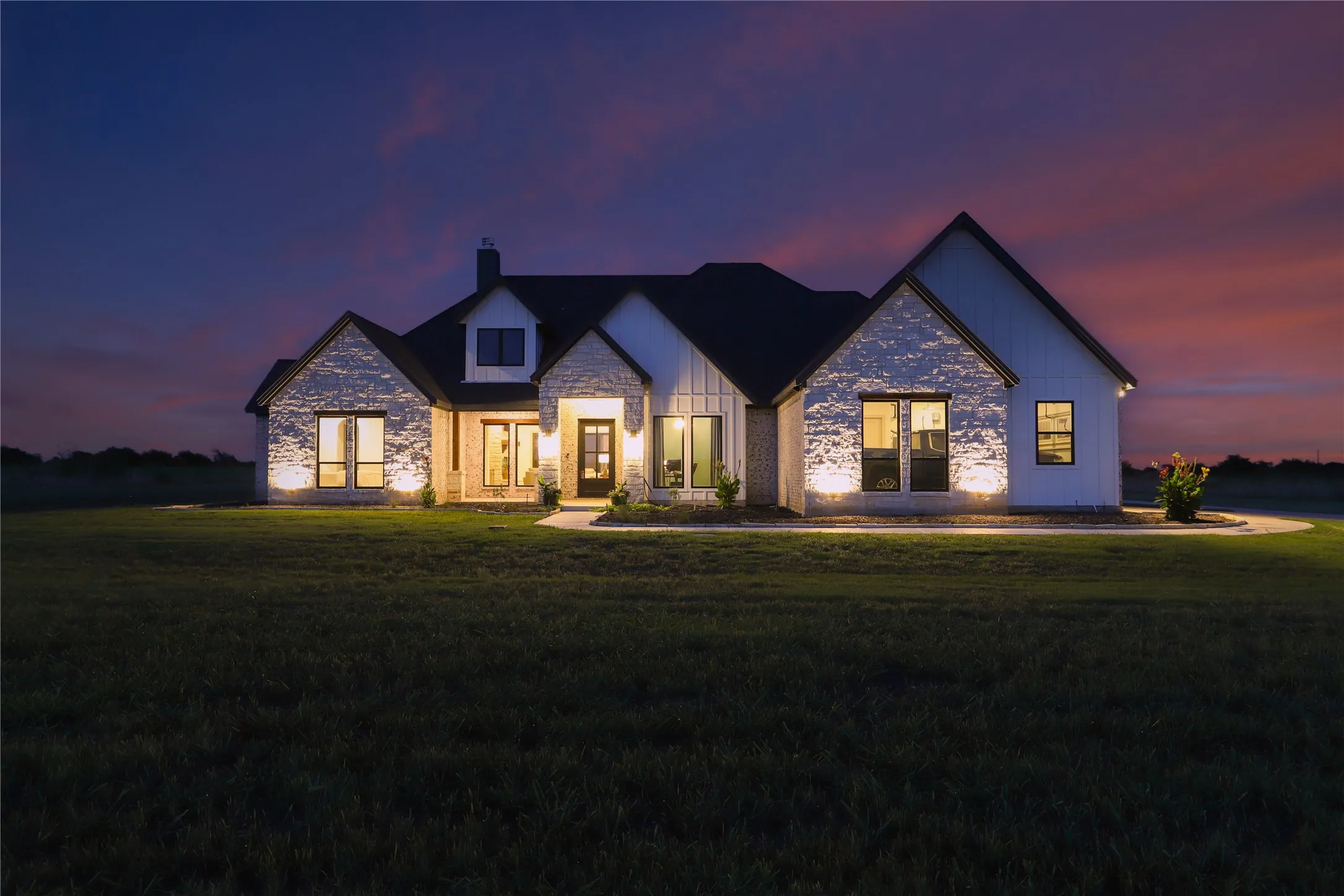 Exterior facade featuring stone and vertical panel siding, dark-framed windows, a covered entry, and integrated exterior lighting