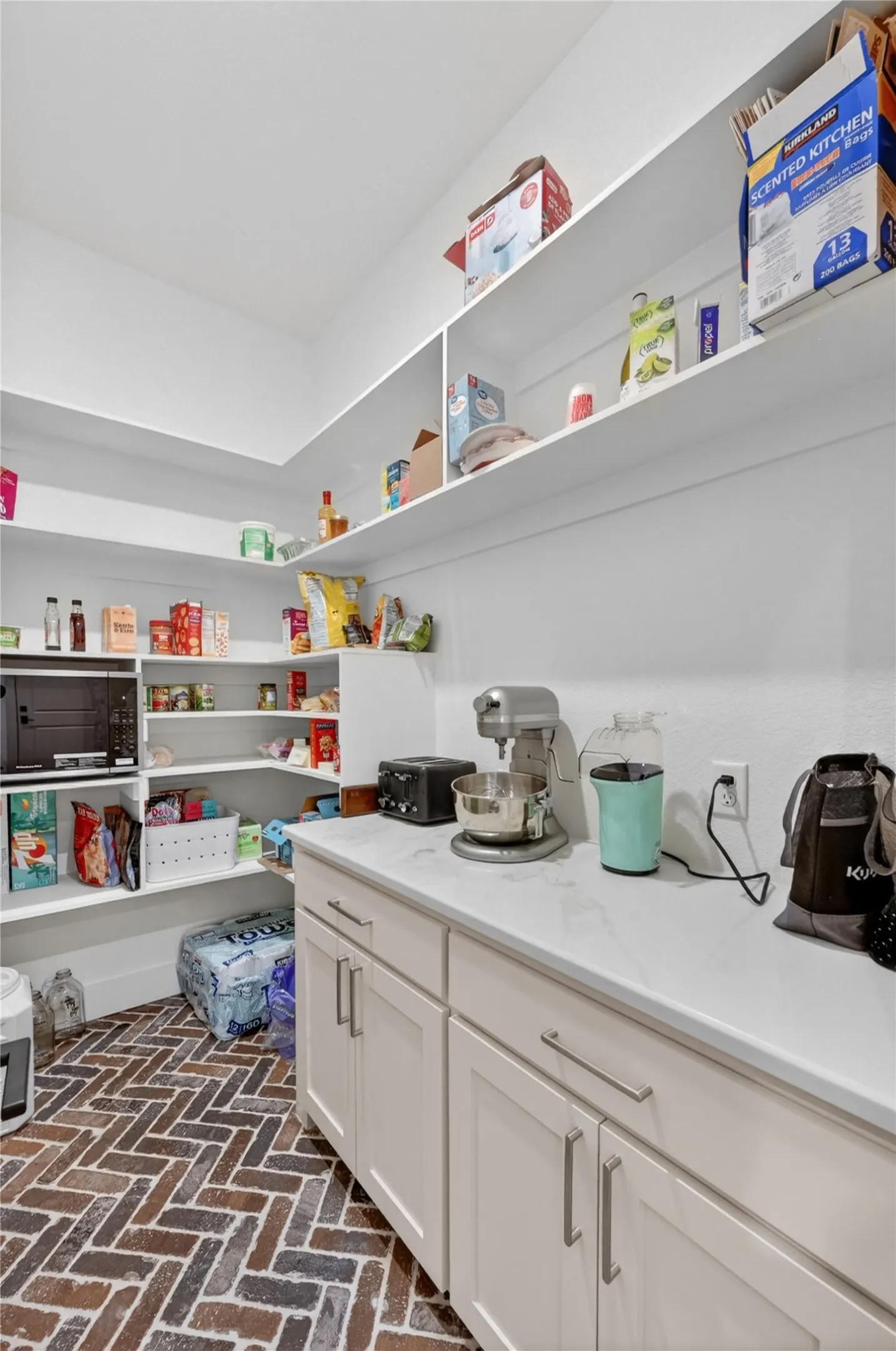 Expansive pantry featuring ample shelving, neutral-toned cabinetry with satin nickel hardware, a durable light-toned countertop, and a herringbone-patterned brick-finish floor