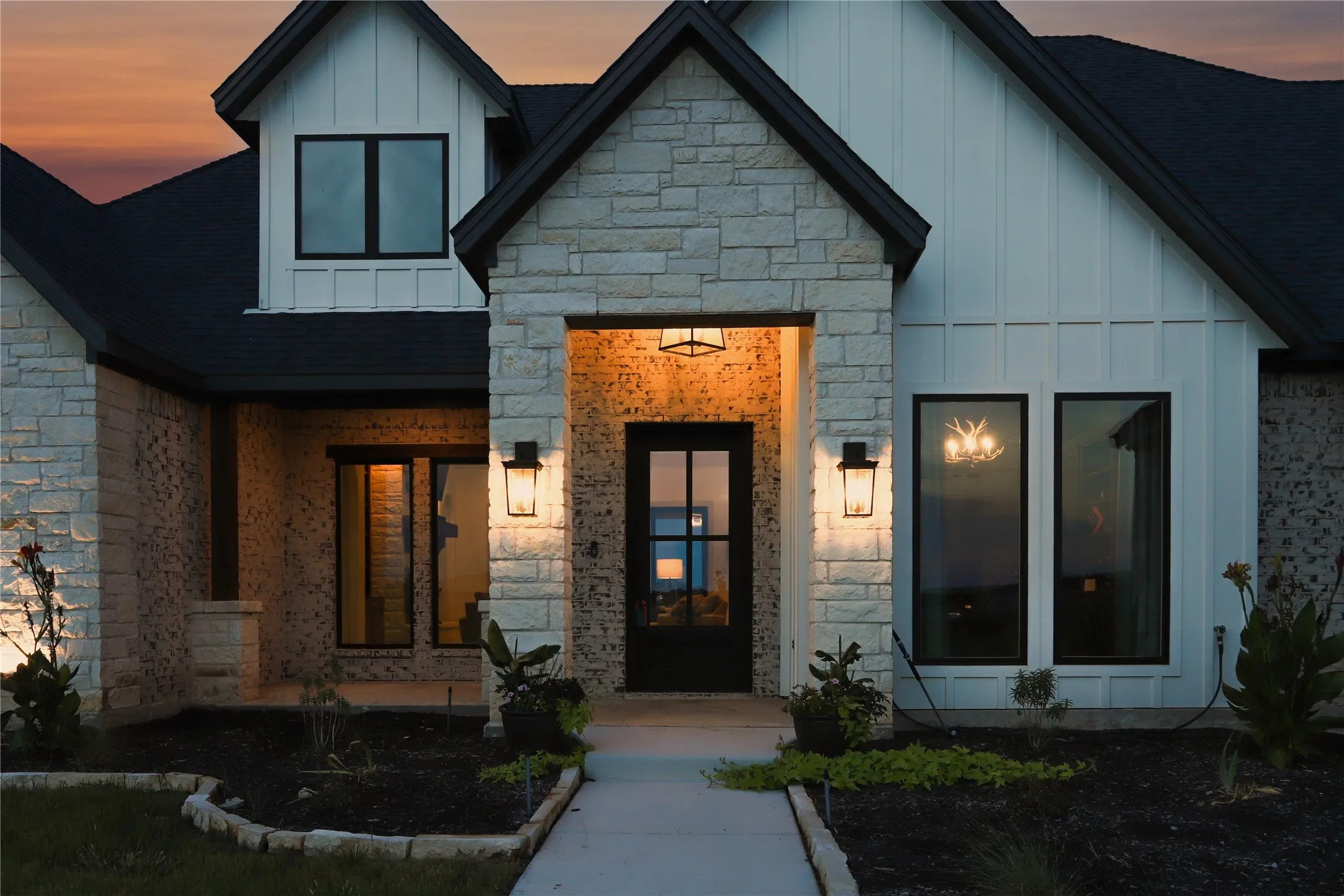 Contemporary home featuring a stone and board-and-batten facade, dark-frame windows, a prominent black entry door with glass inserts, and exterior wall sconces