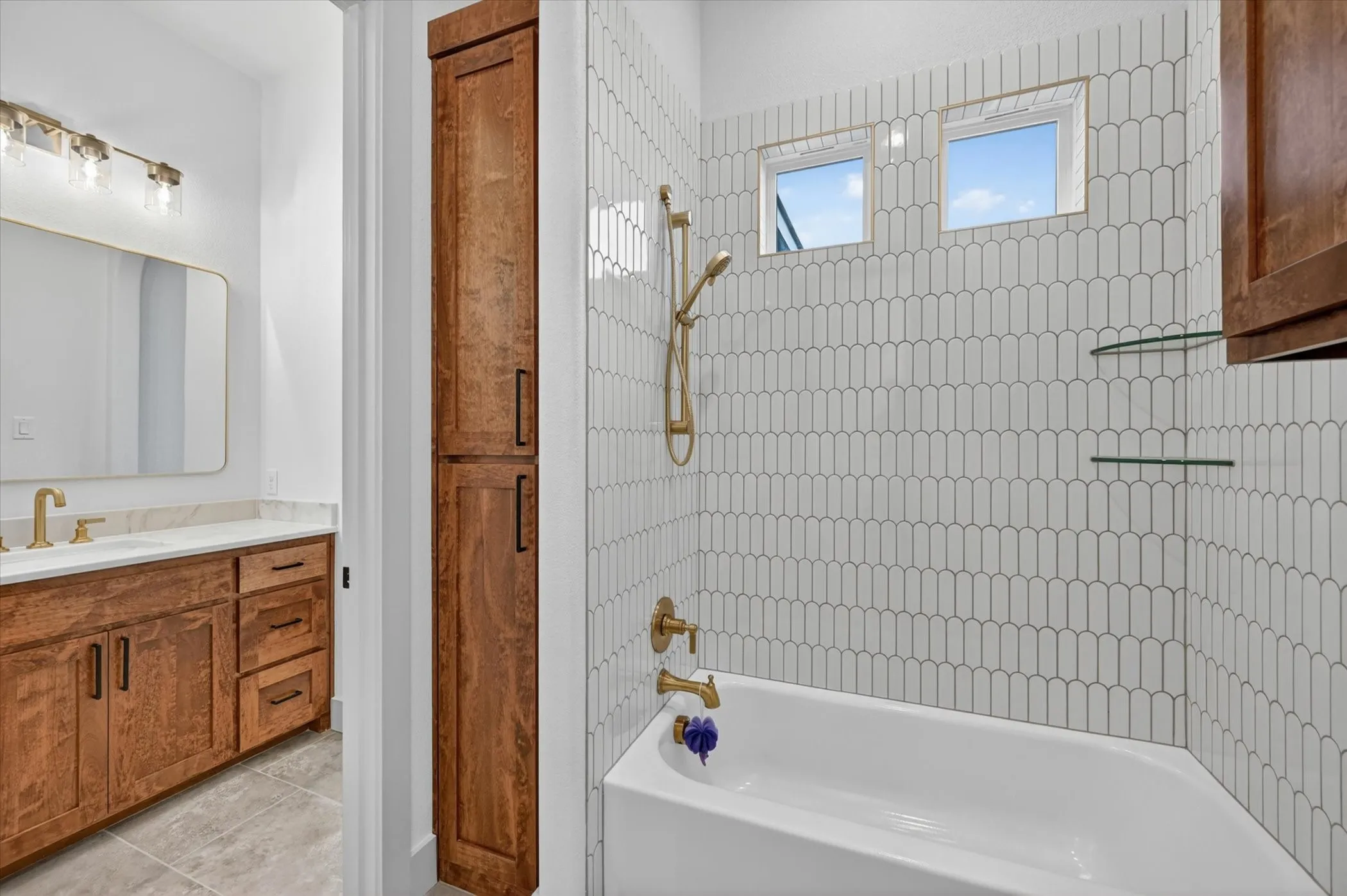 Bathtub with stylized tile surround, dual windows, and brushed gold fixtures