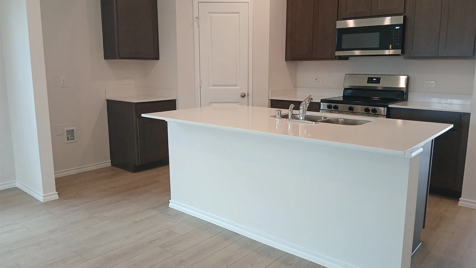 Kitchen featuring a central island with an undermount sink, dark wood-finish cabinetry, light countertops, stainless steel appliances, and wood-finish flooring