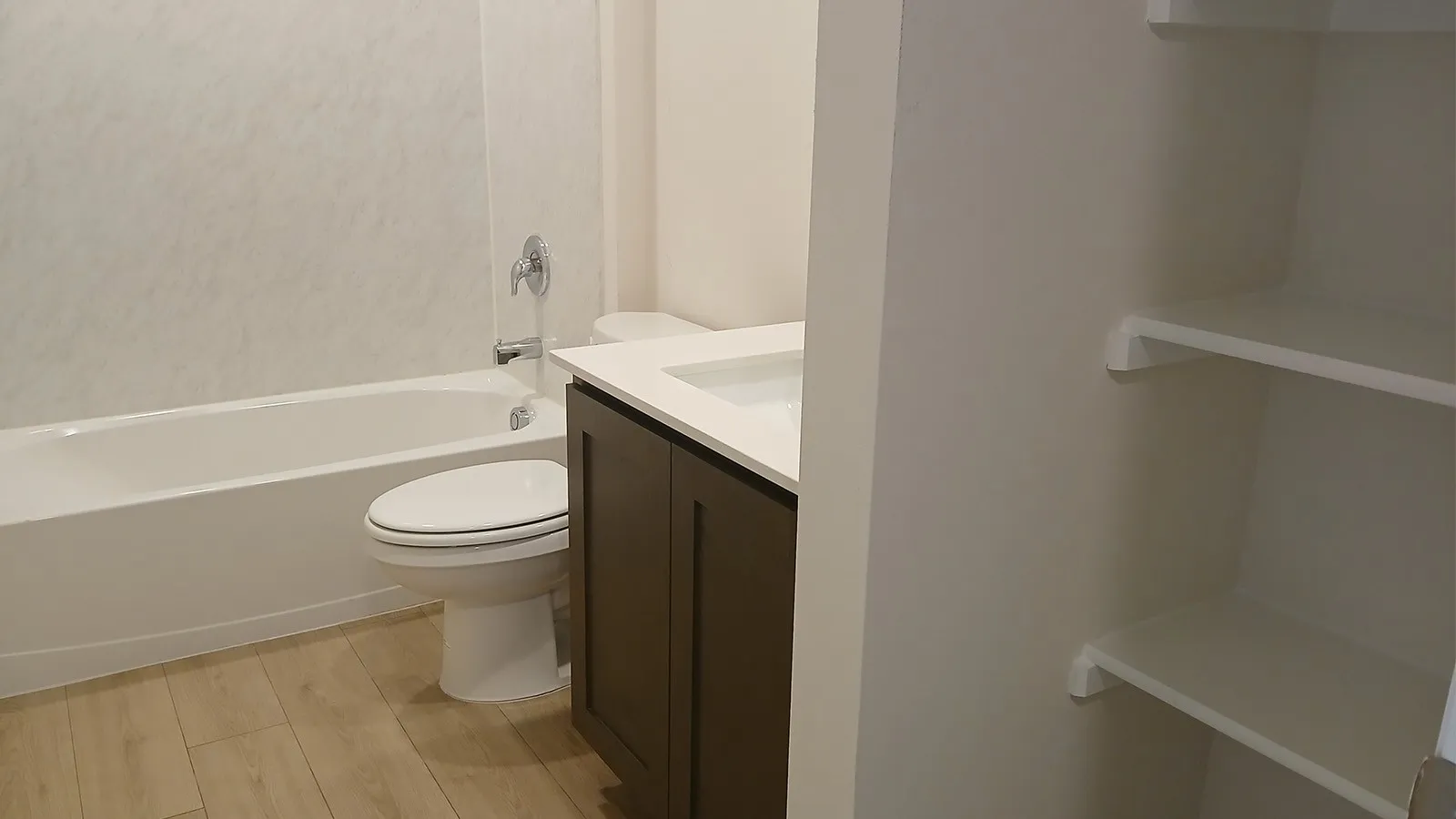 Bathroom featuring a white bathtub with a textured surround, wood-finish flooring, a contemporary toilet, and a vanity with a white countertop and dark cabinetry