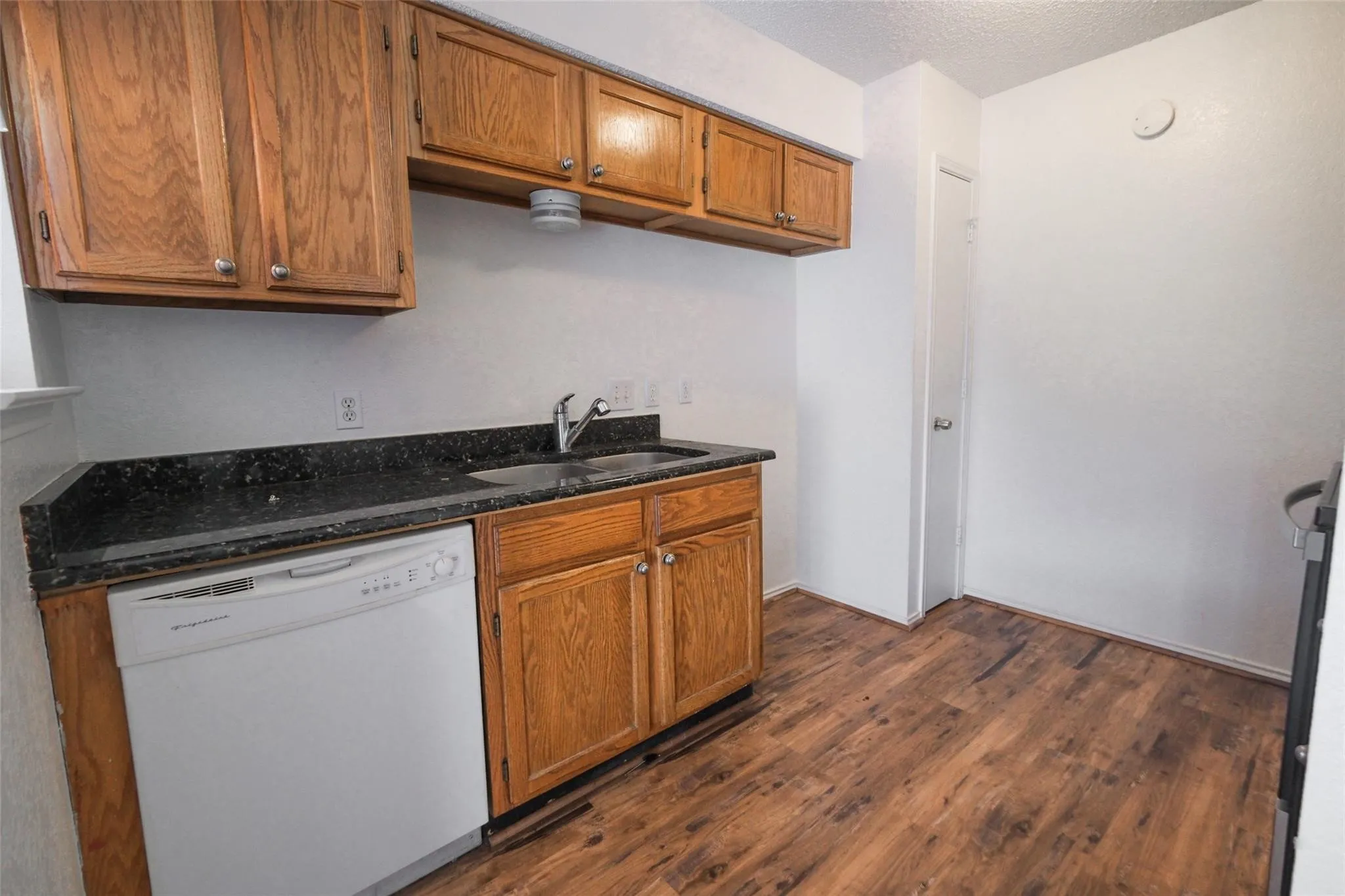 Kitchen featuring wood-finish flooring, dark speckled countertops, wood cabinetry, a stainless steel sink with a chrome faucet, and a white dishwasher