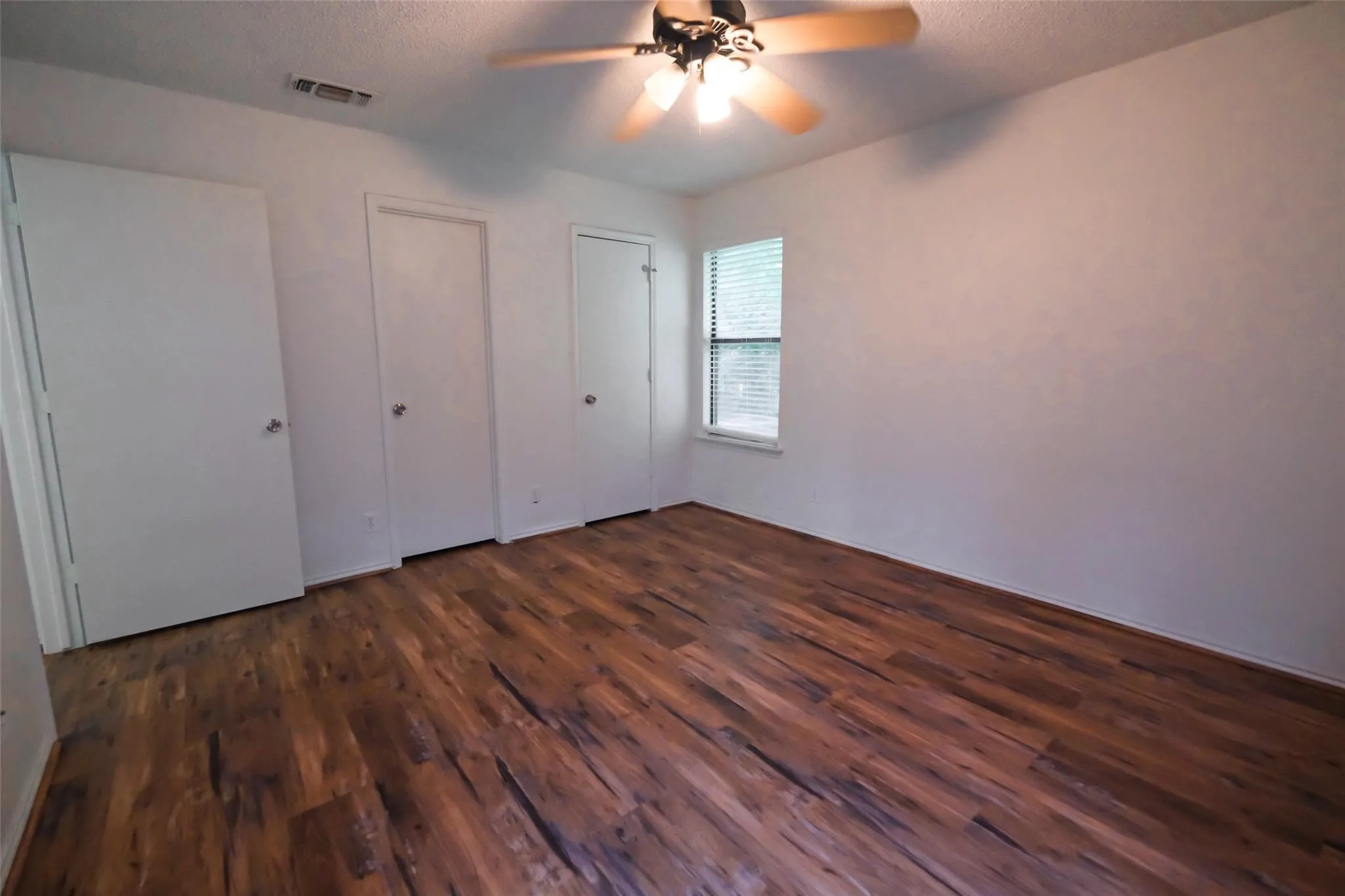 Spacious room featuring wood-finish flooring, a ceiling fan with integrated lighting, a window with blinds, and white interior doors