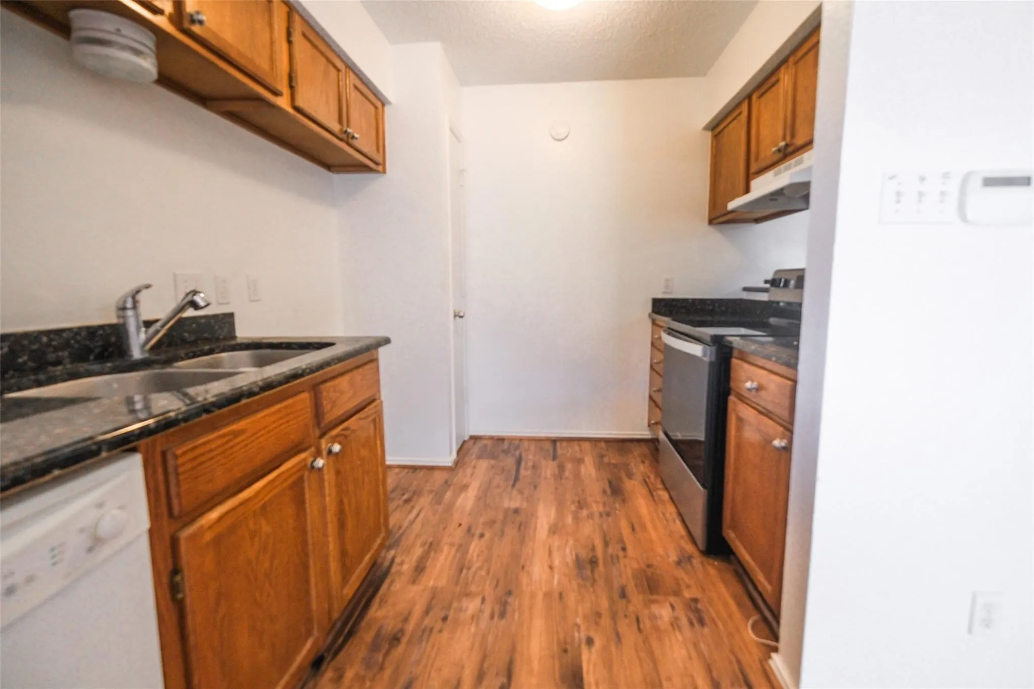 Galley kitchen featuring wood-finish flooring, dark speckled countertops, wood cabinetry, a double basin sink with a chrome faucet, and stainless steel appliances