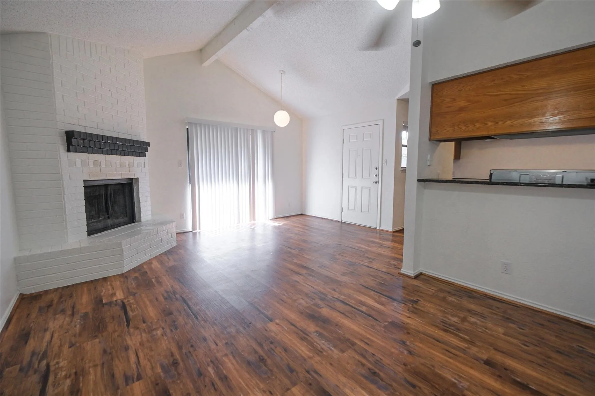 Spacious living area featuring wood-finish flooring, a white brick fireplace with a dark mantel, a high vaulted ceiling with exposed beam, and vertical blinds on a sliding glass door
