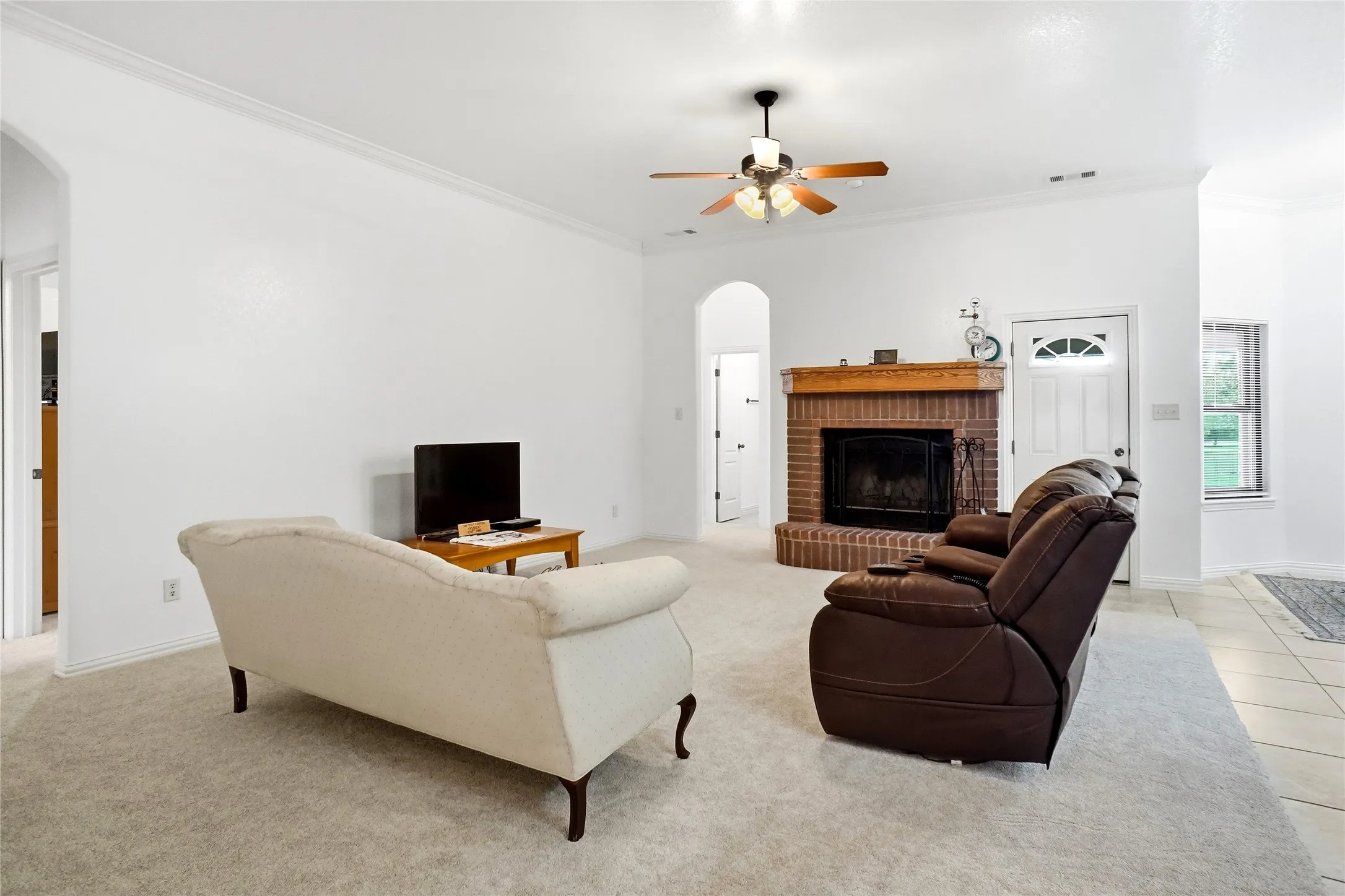 Spacious living area featuring a brick fireplace with a wood mantel, light-toned carpet, a ceiling fan, and crown molding