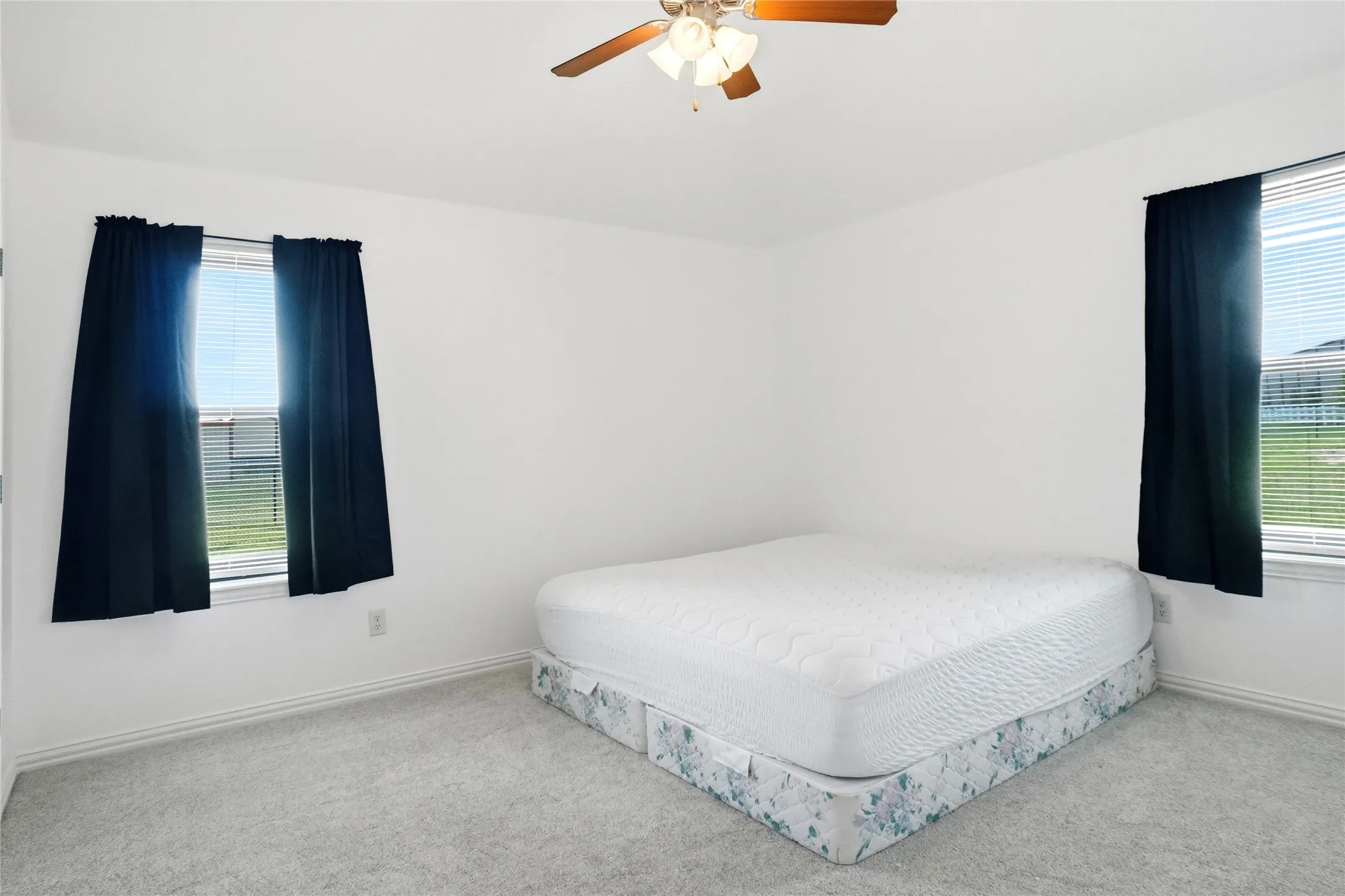 Carpeted room featuring dual windows with blinds and dark curtains, a ceiling fan with light fixture, and white baseboards